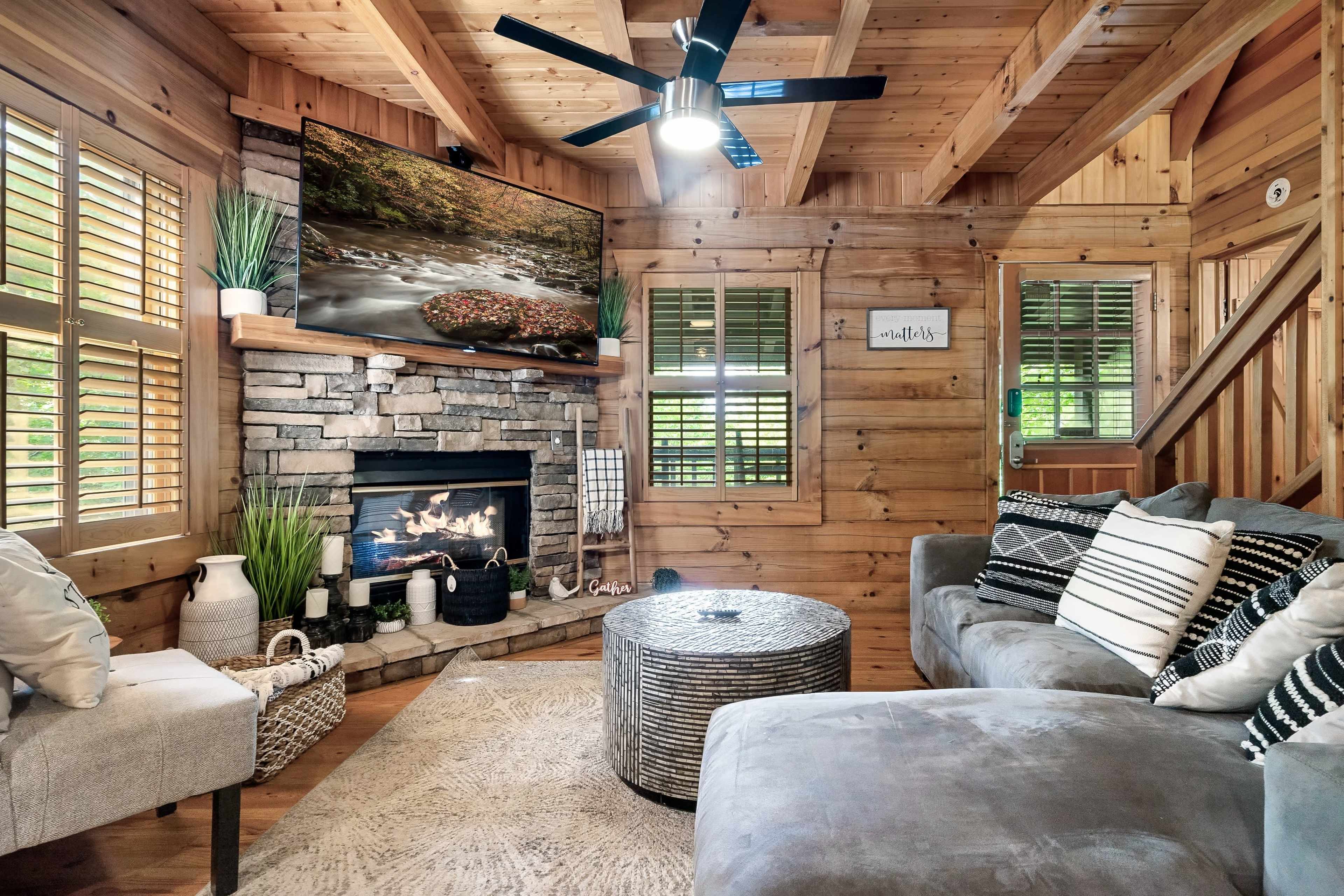 A cozy living room features wooden walls, a stone fireplace, a large television mounted above it, and comfortable seating arranged around a circular table.