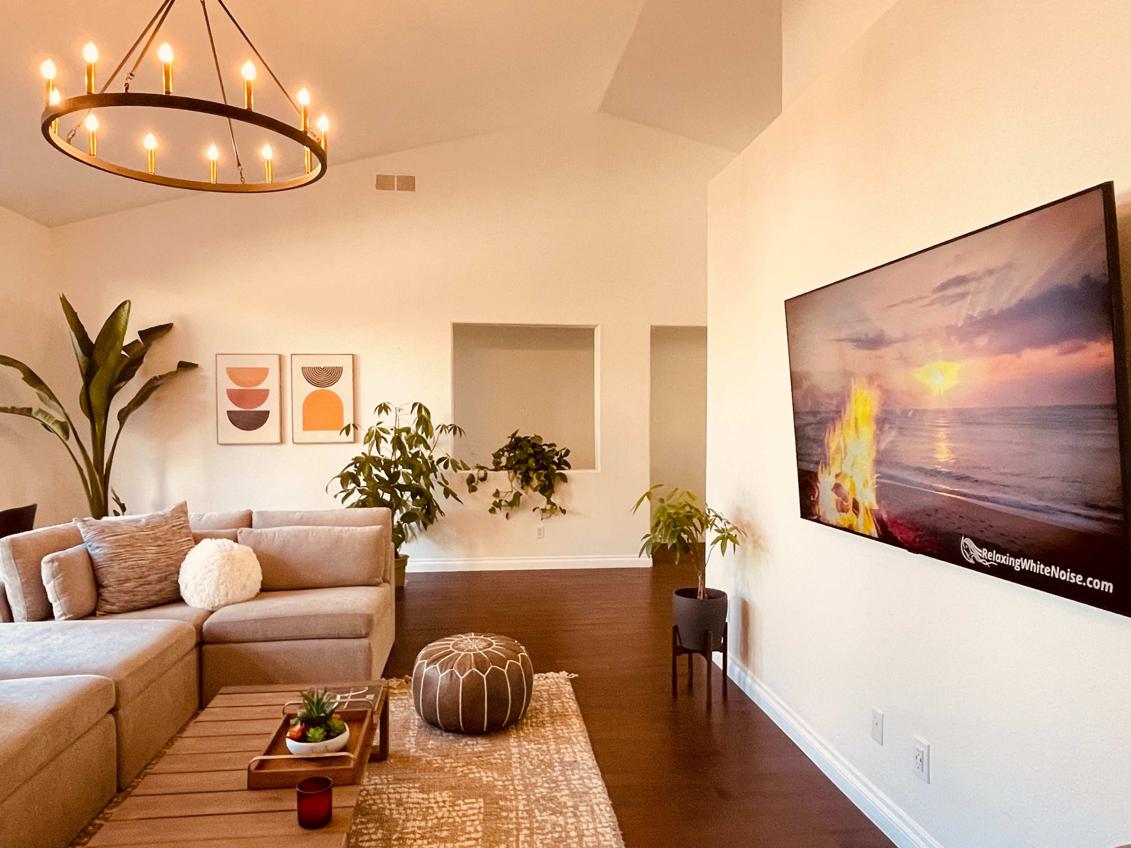 A light-filled living room features a sectional sofa, a television mounted on the wall displaying a beach scene, and various plants arranged throughout the space.
