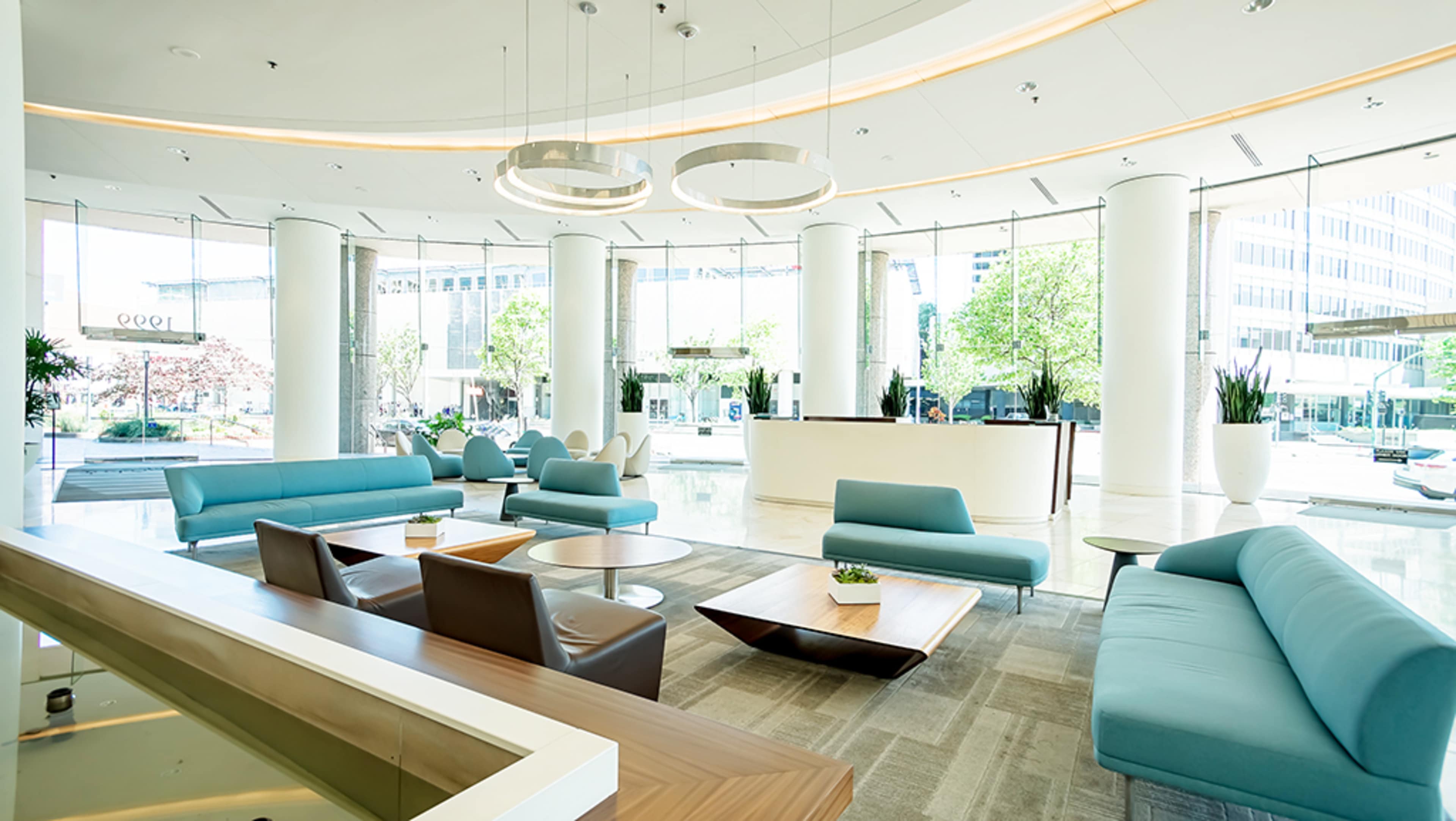 A modern lobby with large windows, white columns, and teal seating arranged around wooden tables.