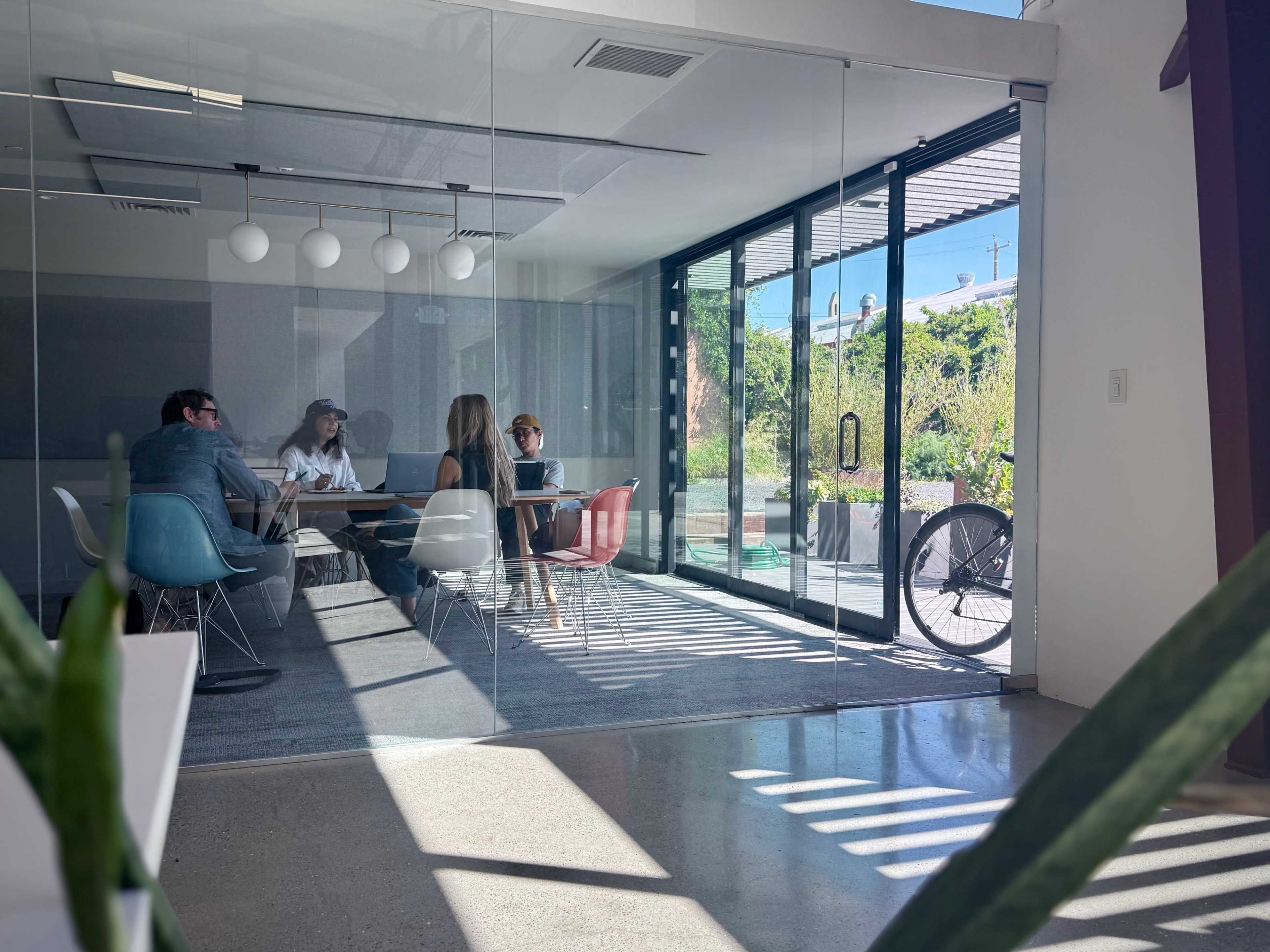 A group of four people is seated around a glass conference table in a bright, modern meeting room with large windows and a bicycle parked outside.