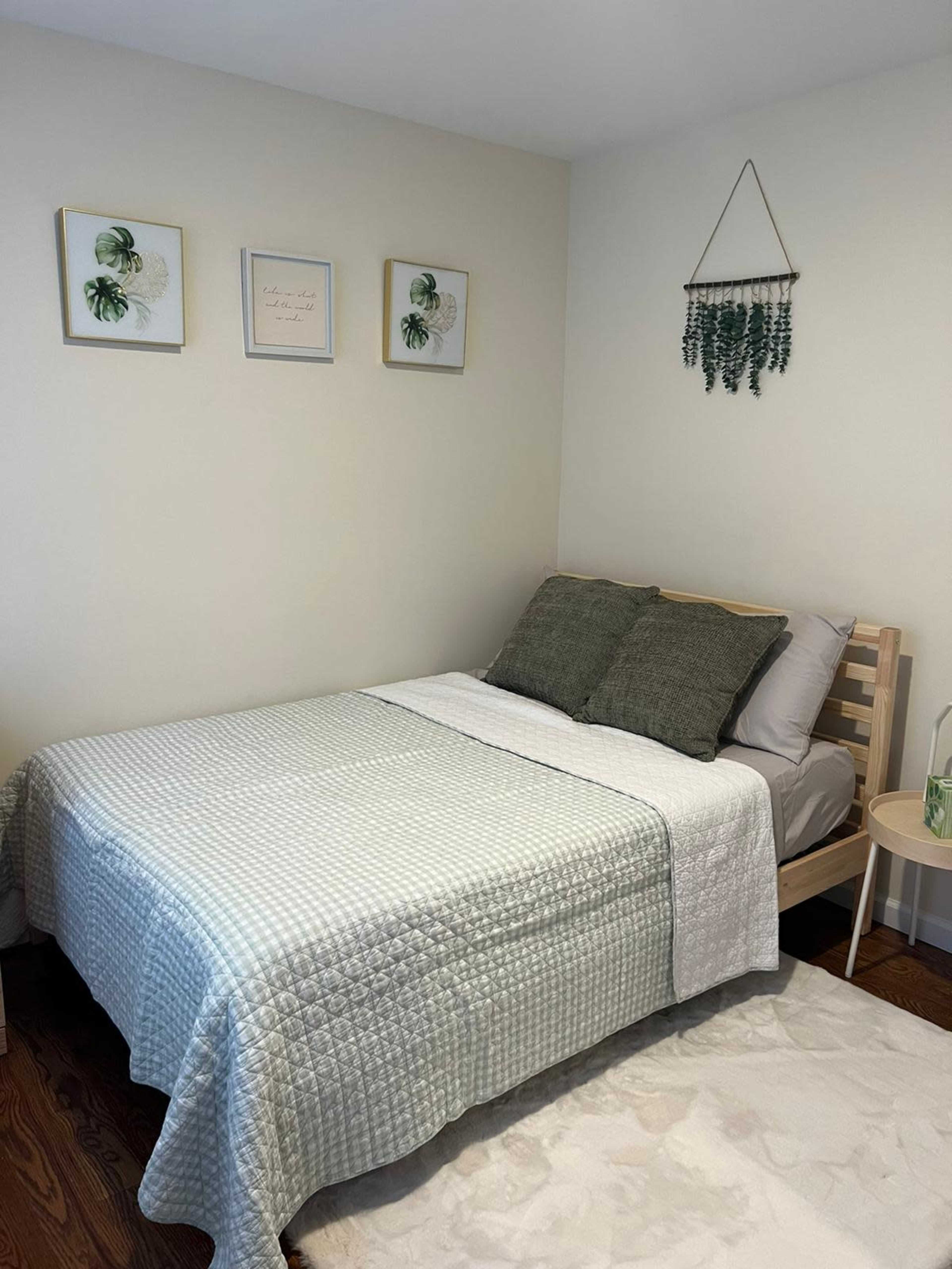 A simple bedroom features a bed with a green and white quilt, framed botanical prints on the wall, and a hanging plant decoration.