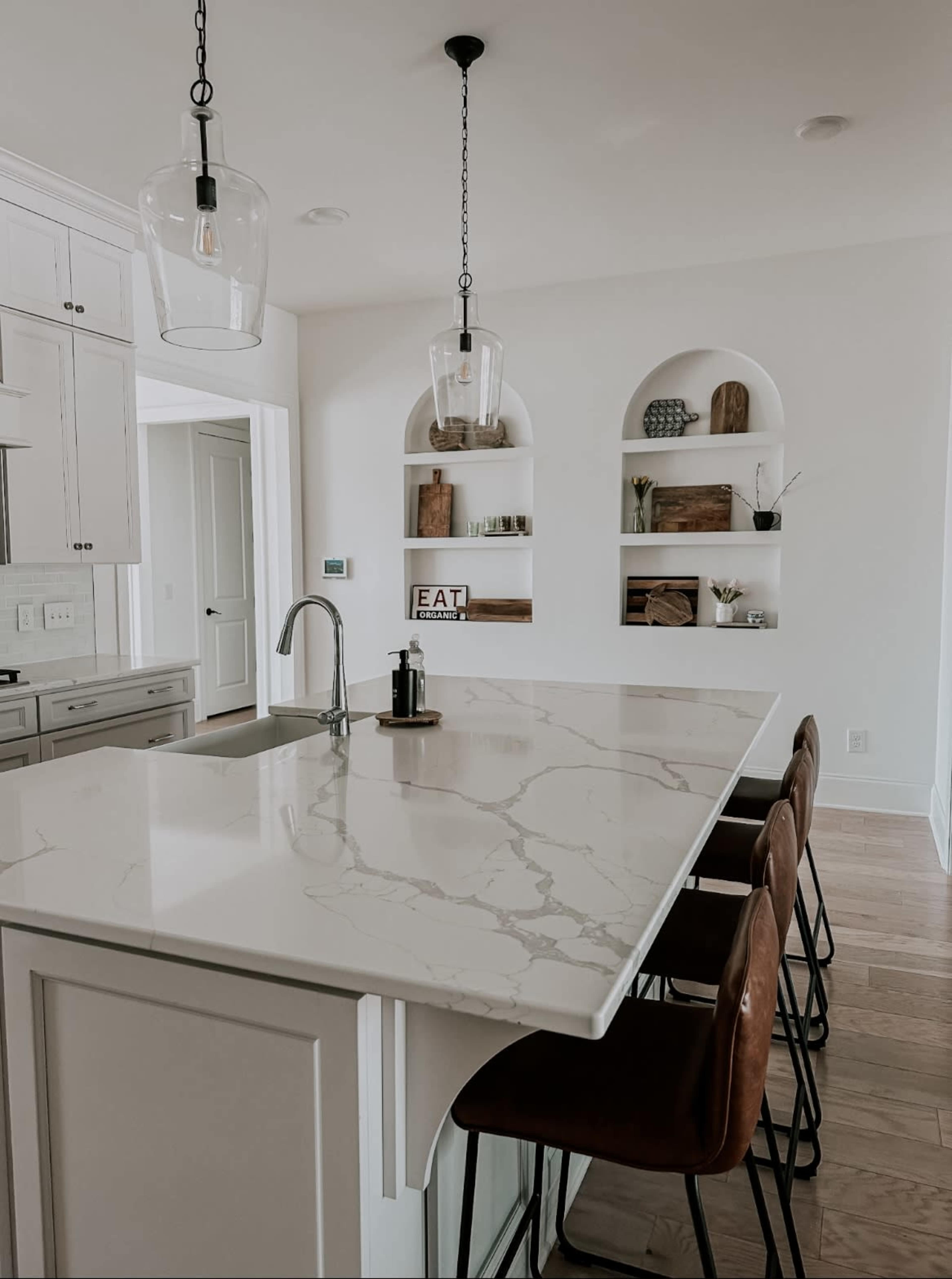 The image shows a modern kitchen with a marble countertop, four brown bar stools, and open shelves displaying decorative items.