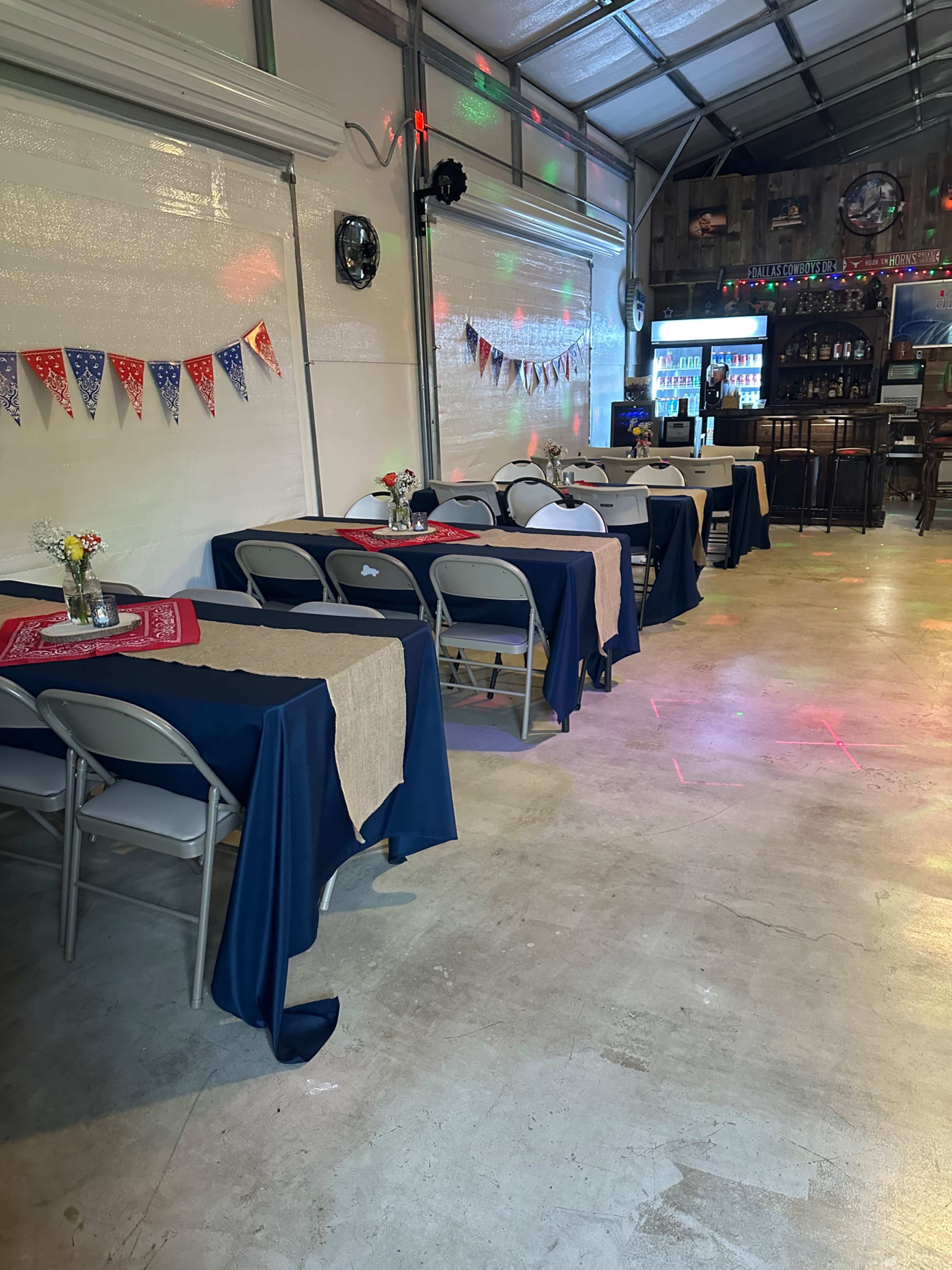 The image shows a decorated indoor space with tables covered in dark blue and beige tablecloths, arranged along the sides, and a bar area in the background illuminated with colorful lights.