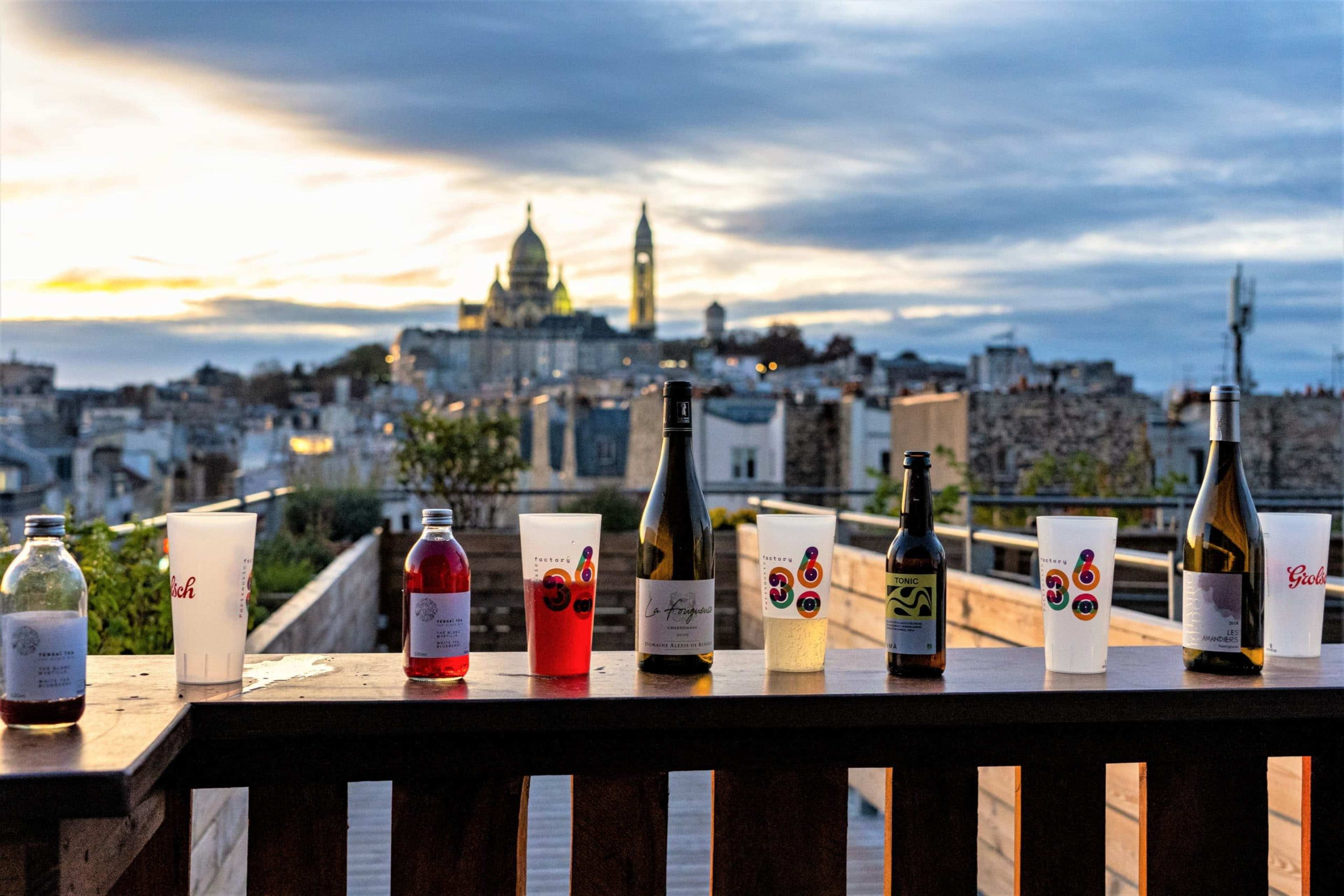 Eight bottles and glasses of various beverages are lined up on a wooden railing with a view of a cityscape at sunset, featuring prominent church domes in the background.