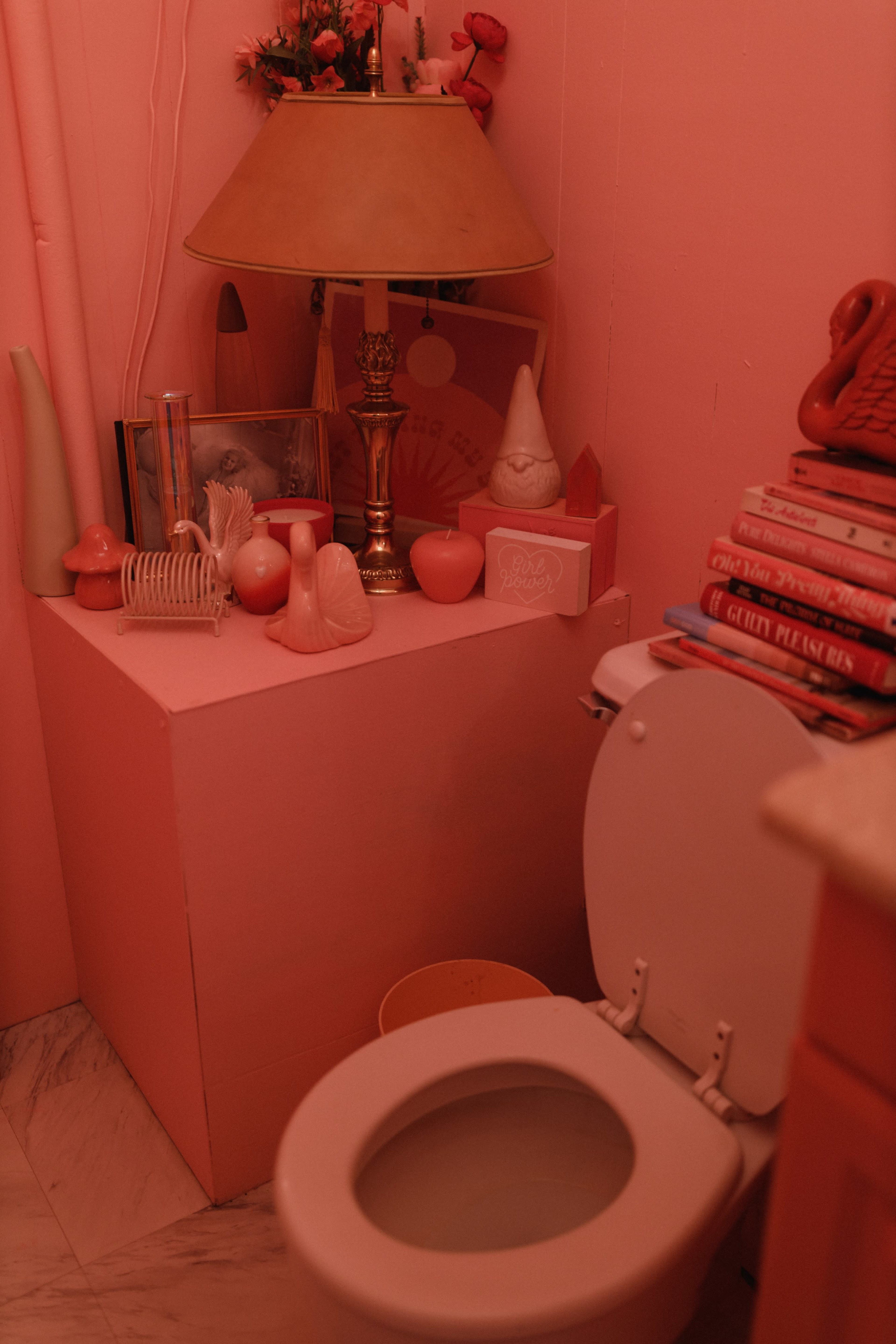 A pink-themed bathroom features a toilet beside a decorative shelf filled with various pink items, including a lamp and books.