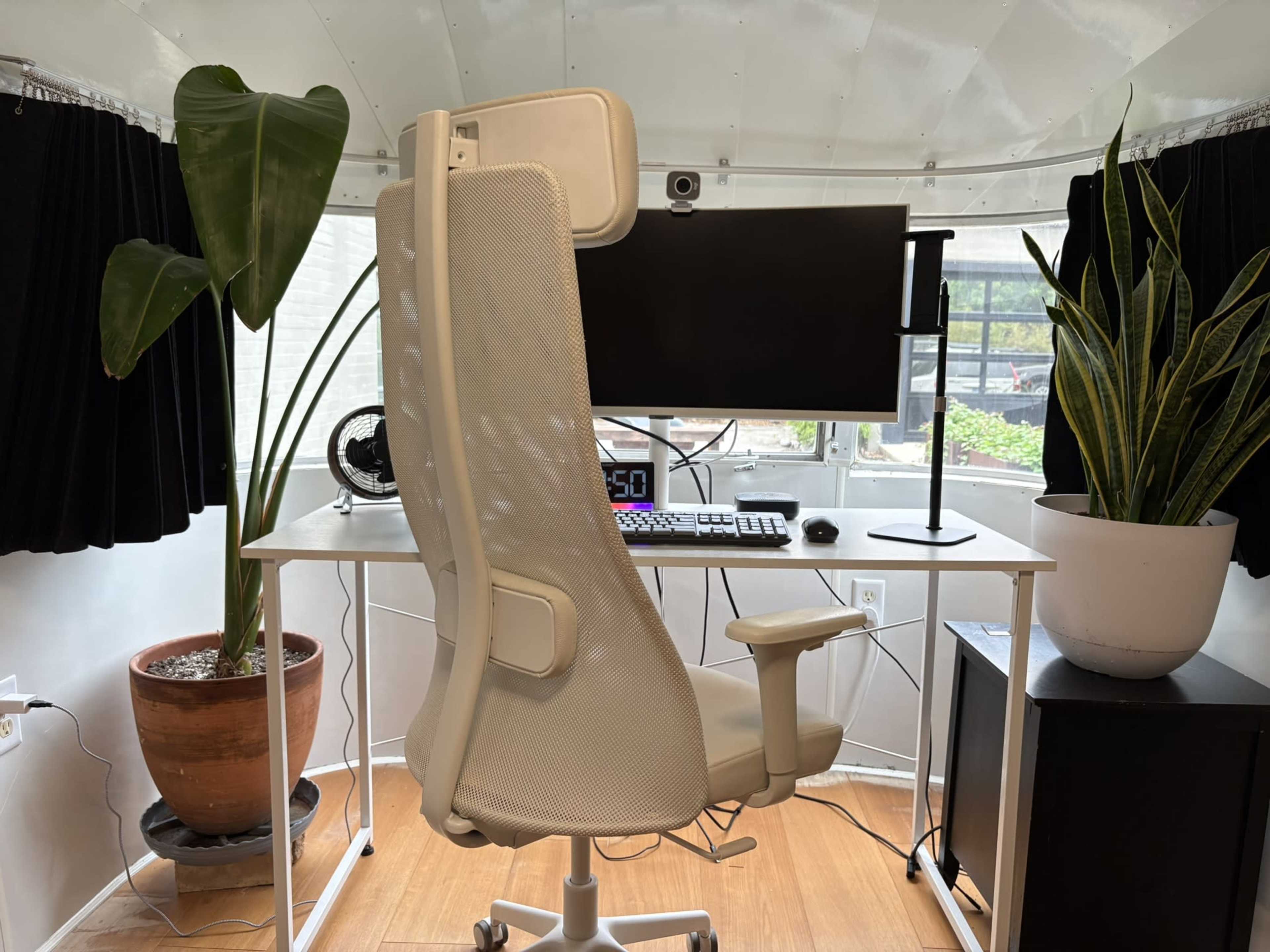 The image shows a modern workspace with a white ergonomic chair facing a large computer monitor on a desk, flanked by two potted plants.