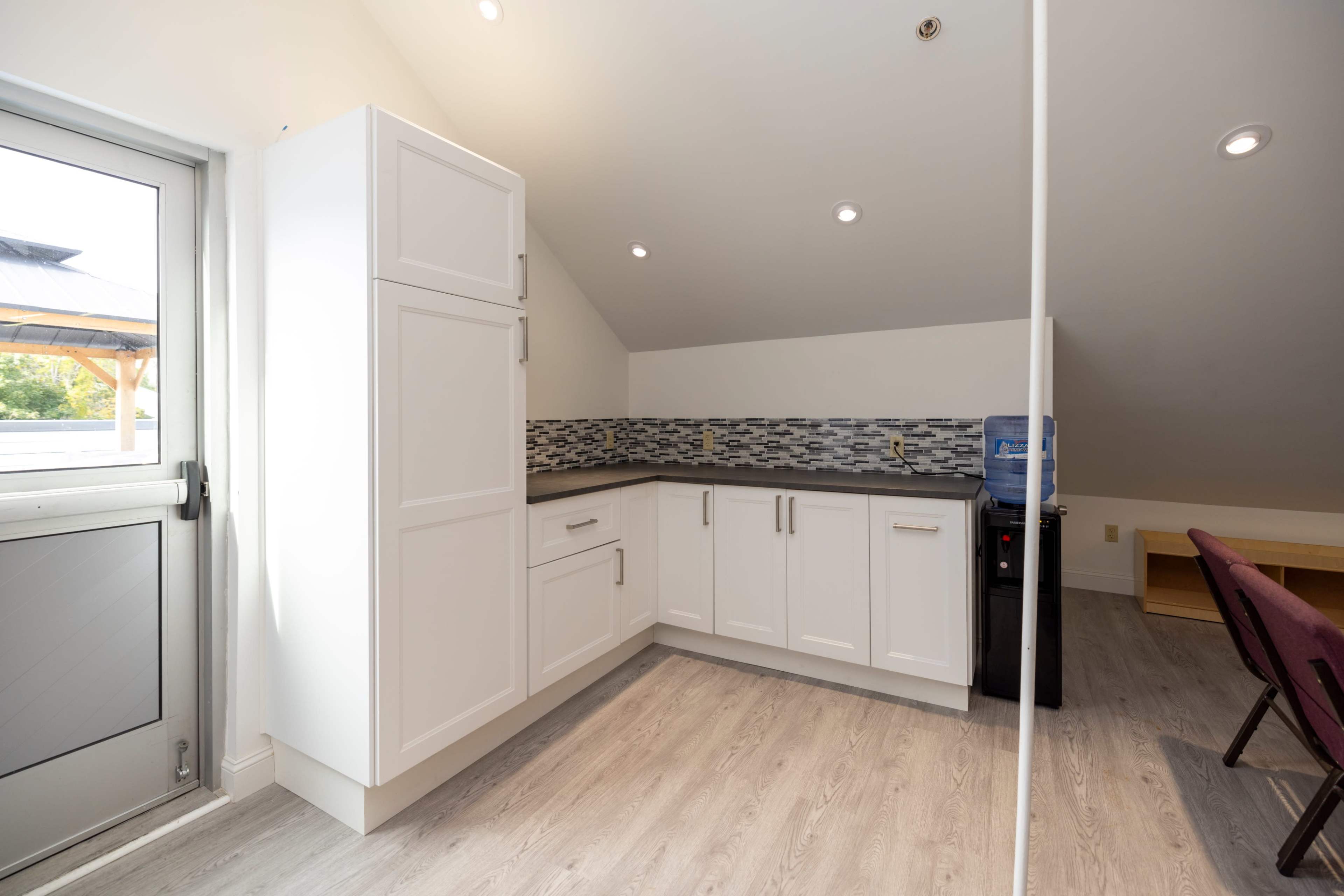 A modern kitchen area with white cabinetry, a gray backsplash, and a water cooler, illuminated by ceiling lights.