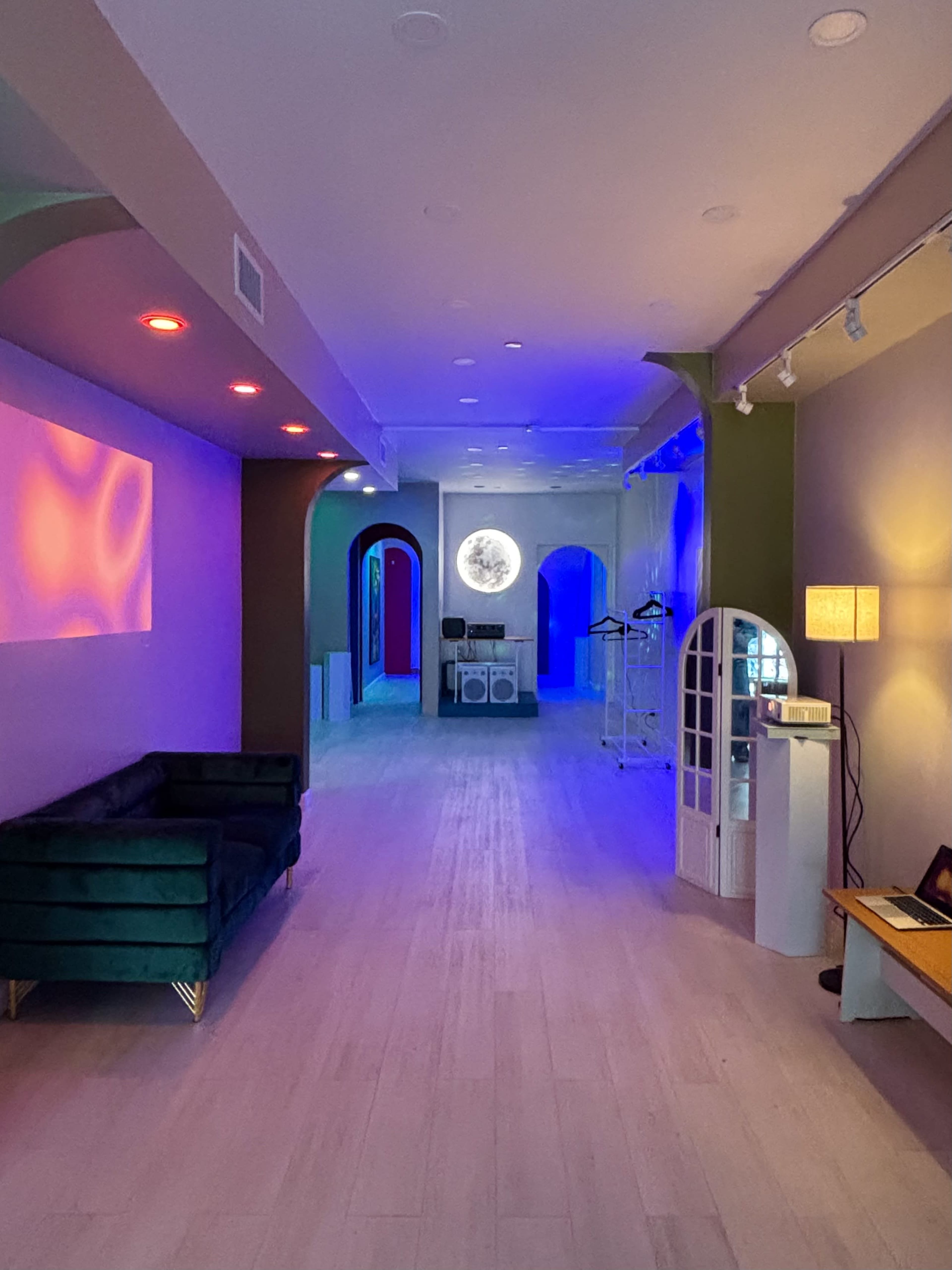 The image shows a modern interior space with colorful lighting, featuring a green velvet sofa on the left and archway entrances leading to different areas.
