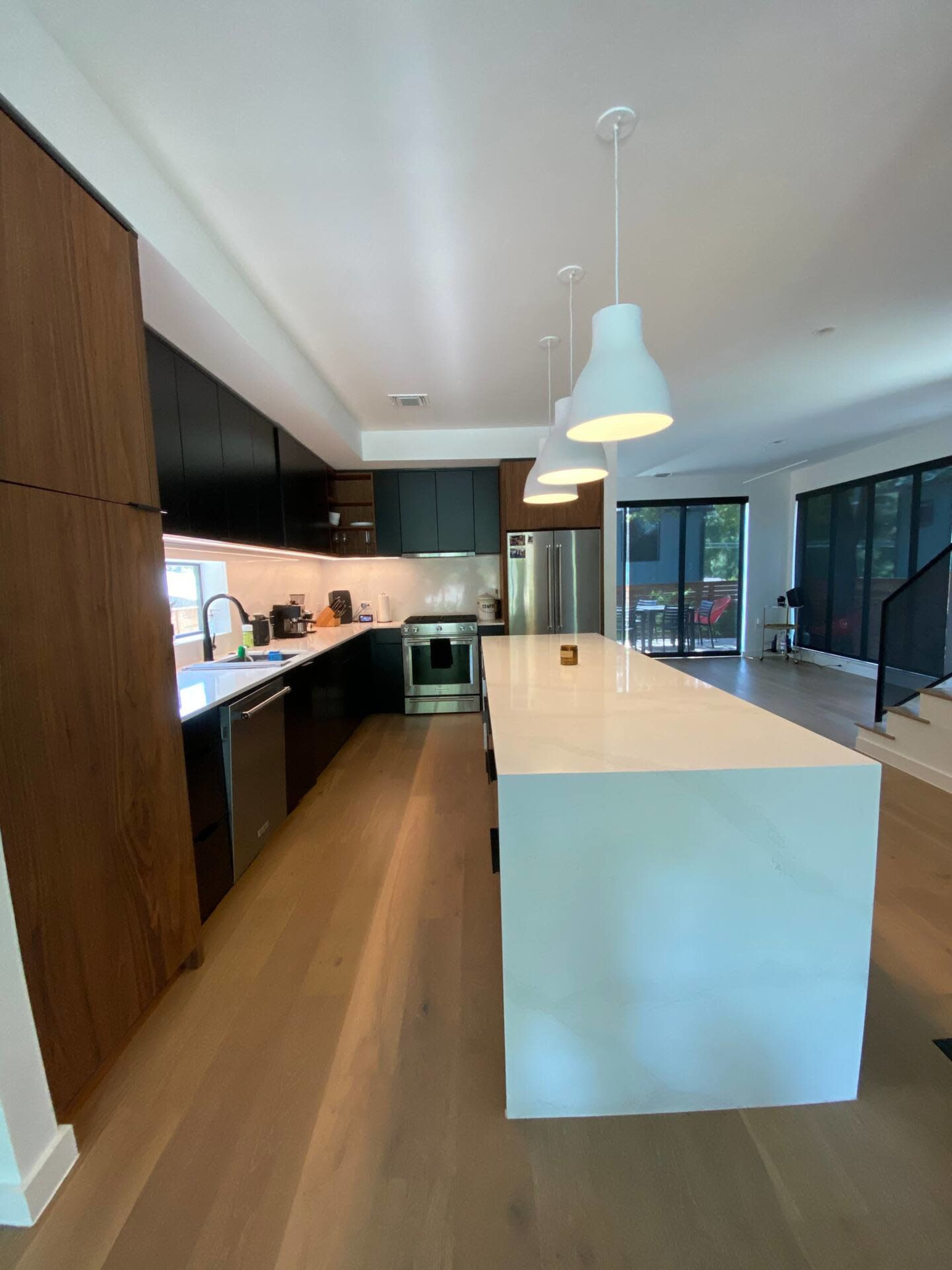 A modern kitchen features dark cabinetry, a white countertop island with pendant lighting, and large windows offering natural light.