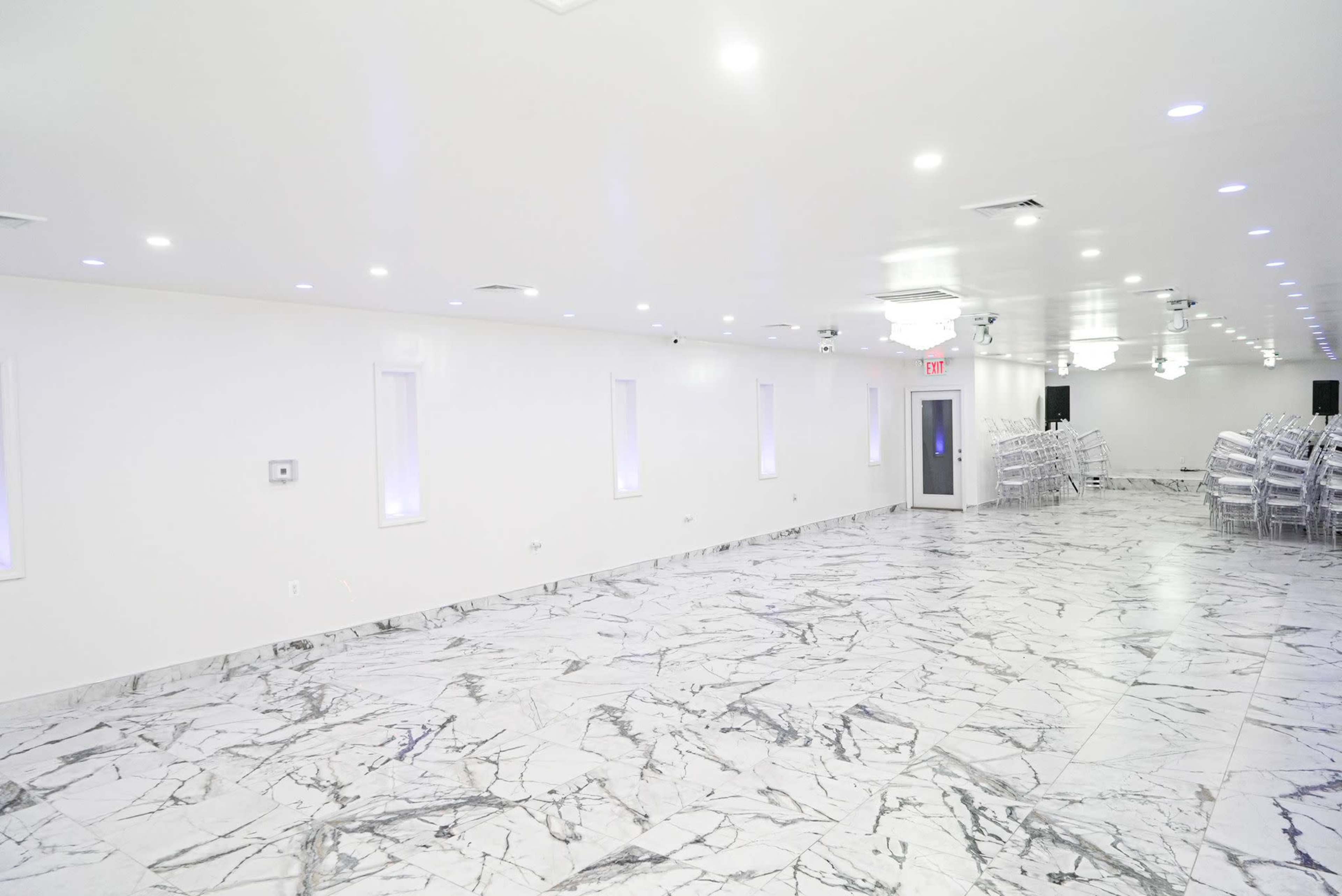 The image shows a spacious, empty hall with white walls, marble flooring, and rows of transparent chairs arranged against one side.