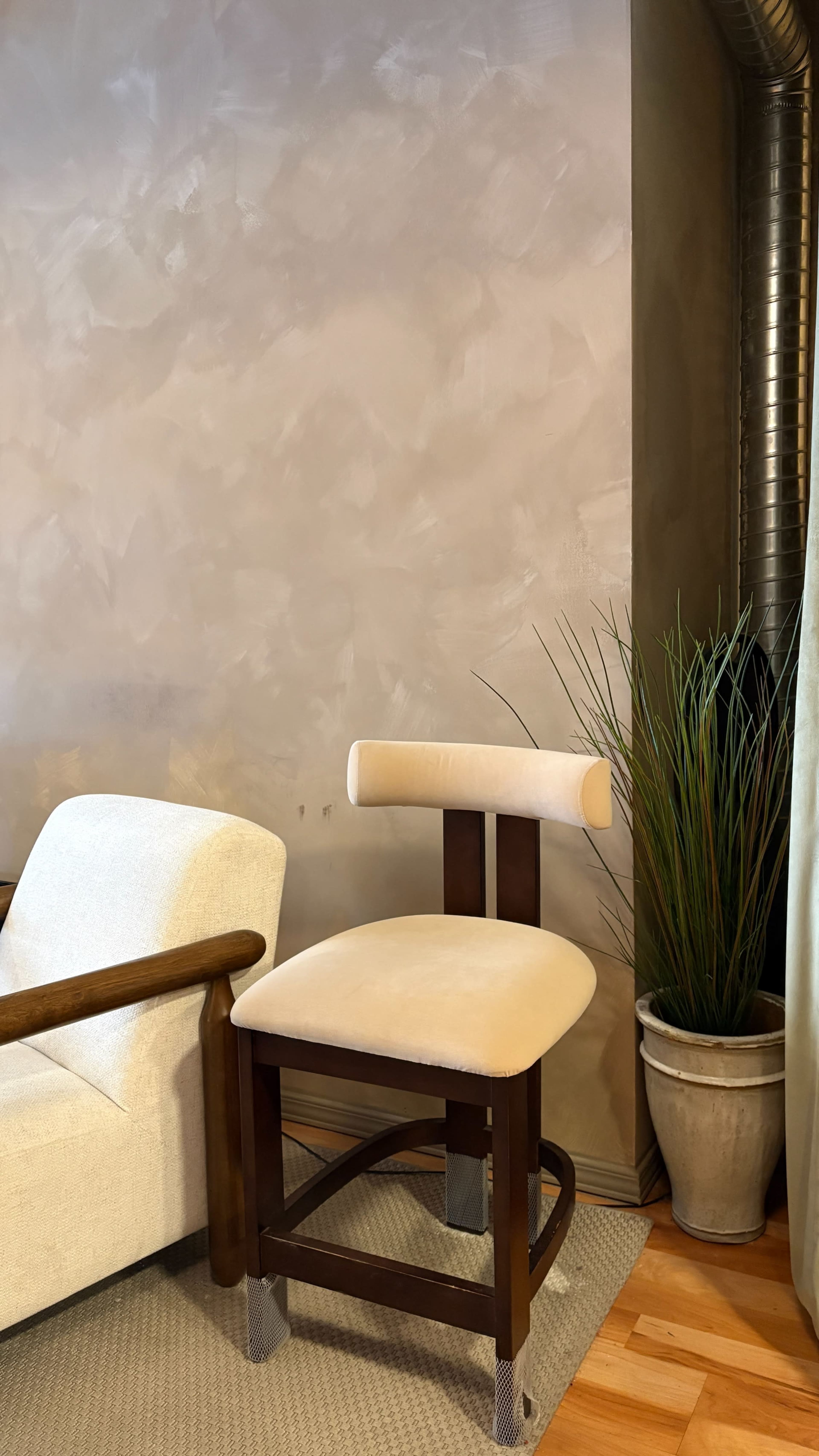 A wooden chair with a beige cushioned seat is positioned next to a light-colored armchair against a textured wall, with a potted plant in the corner.