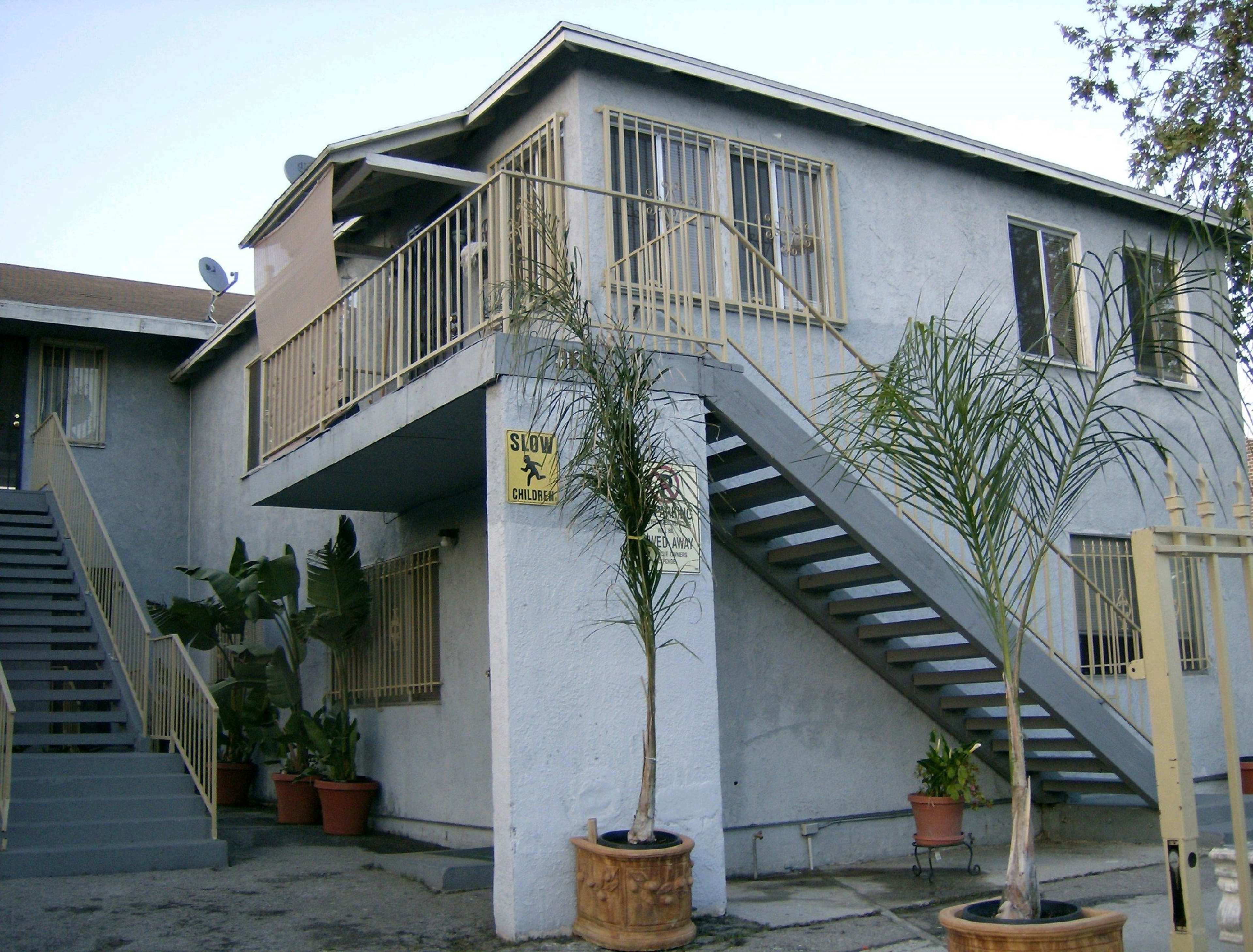 The image shows a two-story apartment building with a gray exterior, featuring a staircase on the left and potted palm plants at the entrance.