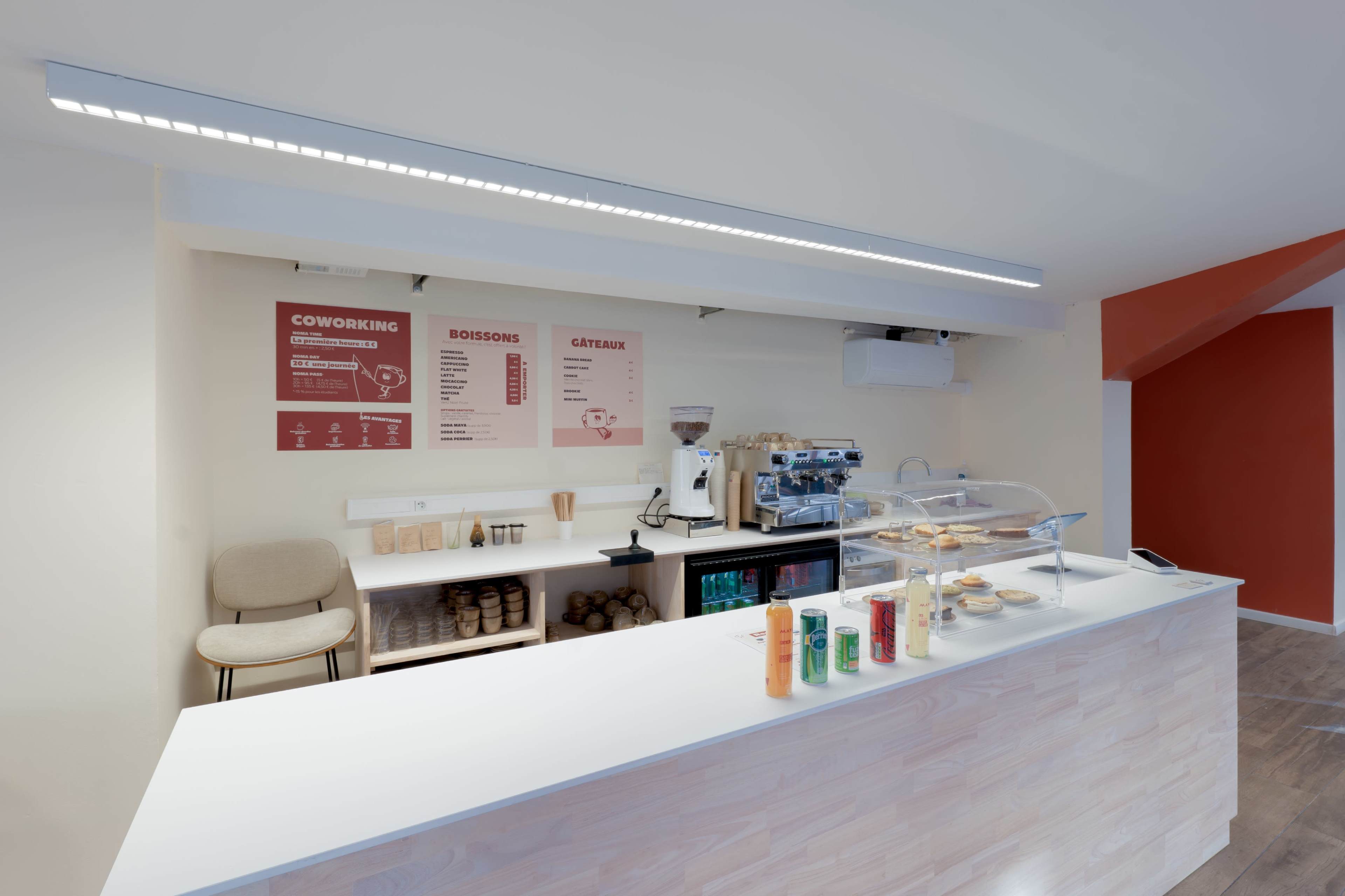 The image shows a modern coffee shop counter with a display case of pastries, coffee machines, and a menu on the wall listing coworking and drink options.