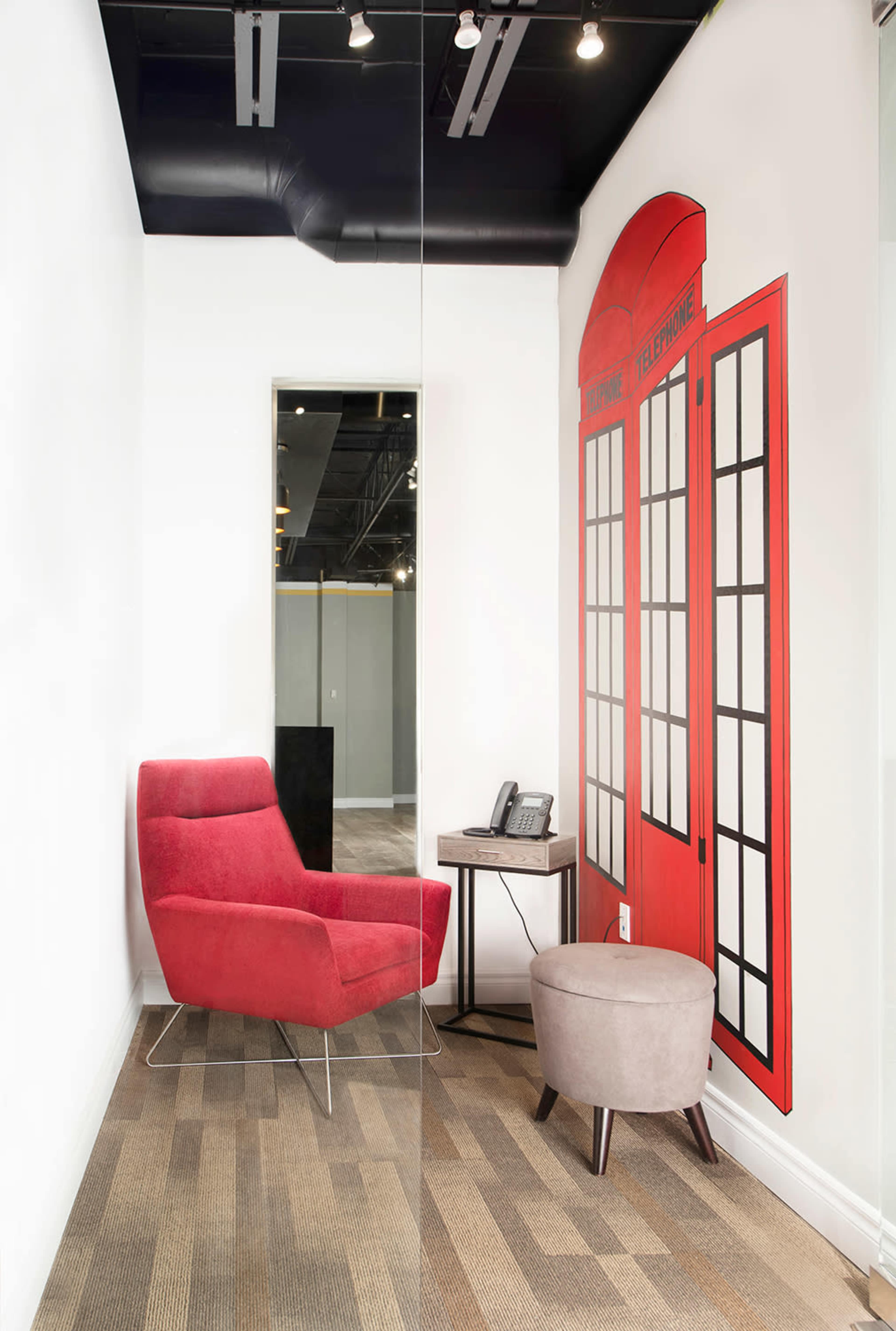A small office space featuring a red chair, a round gray stool, a wooden side table with a telephone, and a wall decorated with a red telephone booth graphic.
