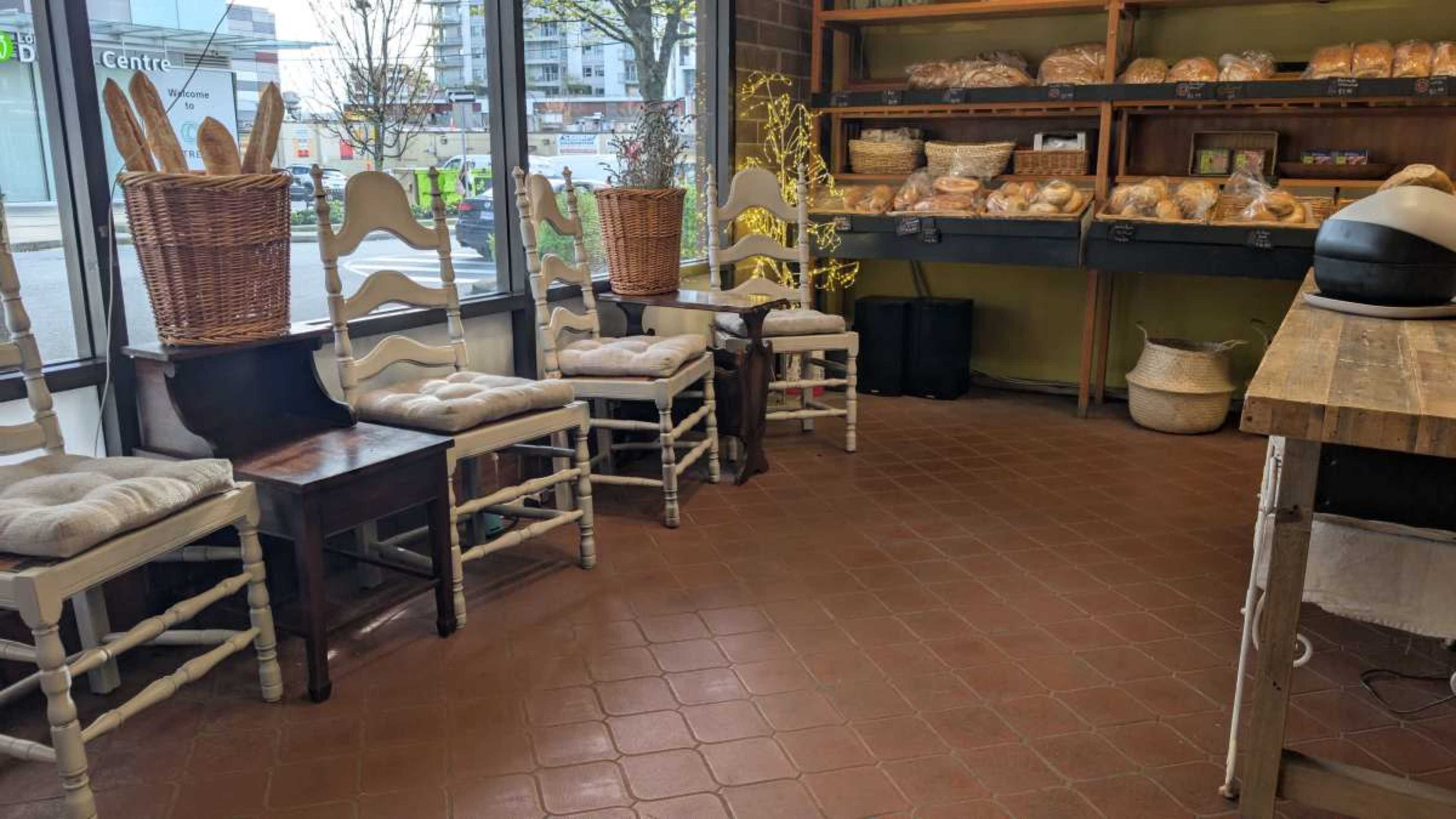 The image shows a cozy bakery interior with wooden chairs, baskets of bread on shelves, and a warm, inviting atmosphere.