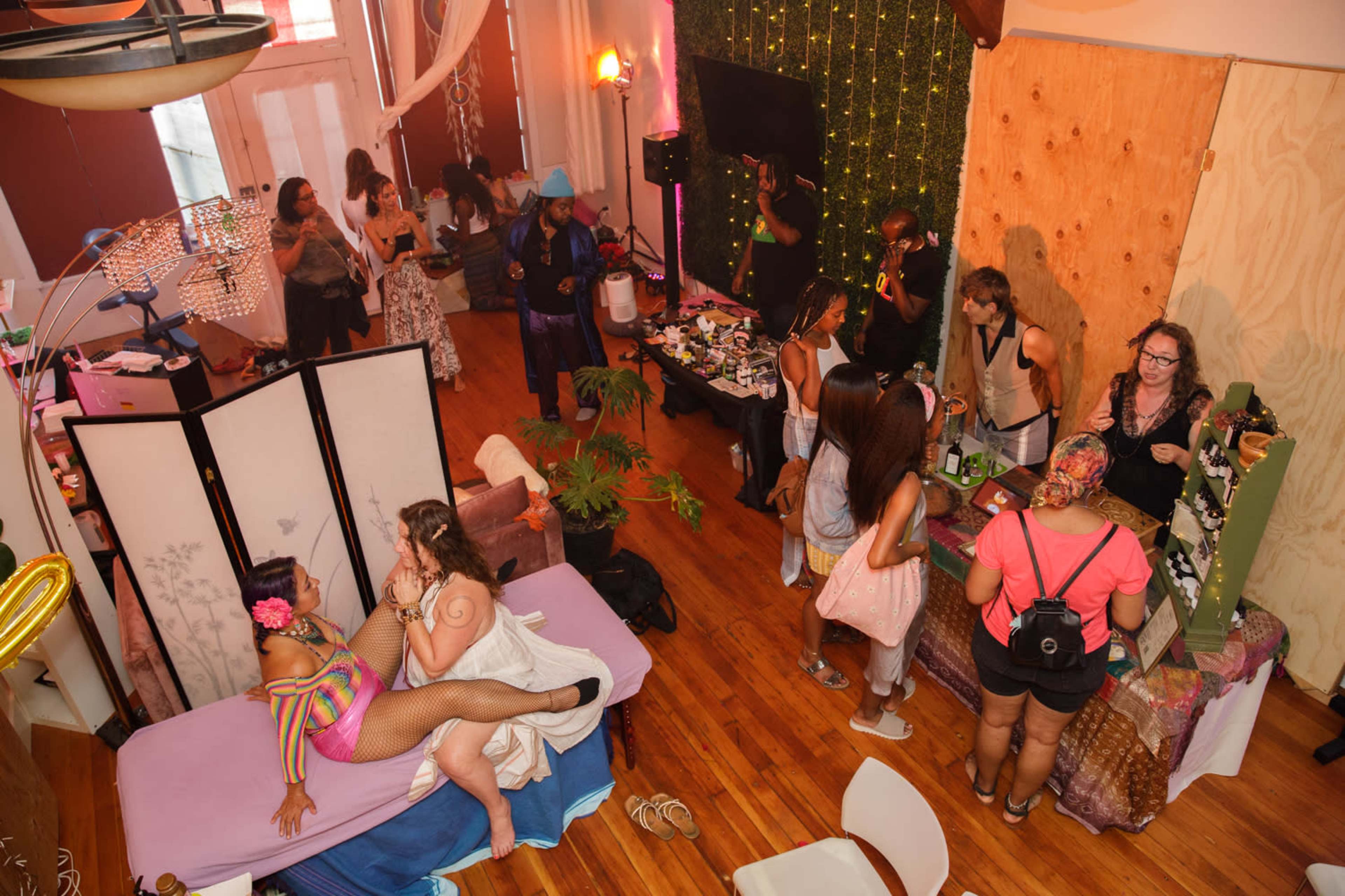 The image shows a lively indoor event space with diverse attendees engaged in various activities, including a performance area and a vendor setup.
