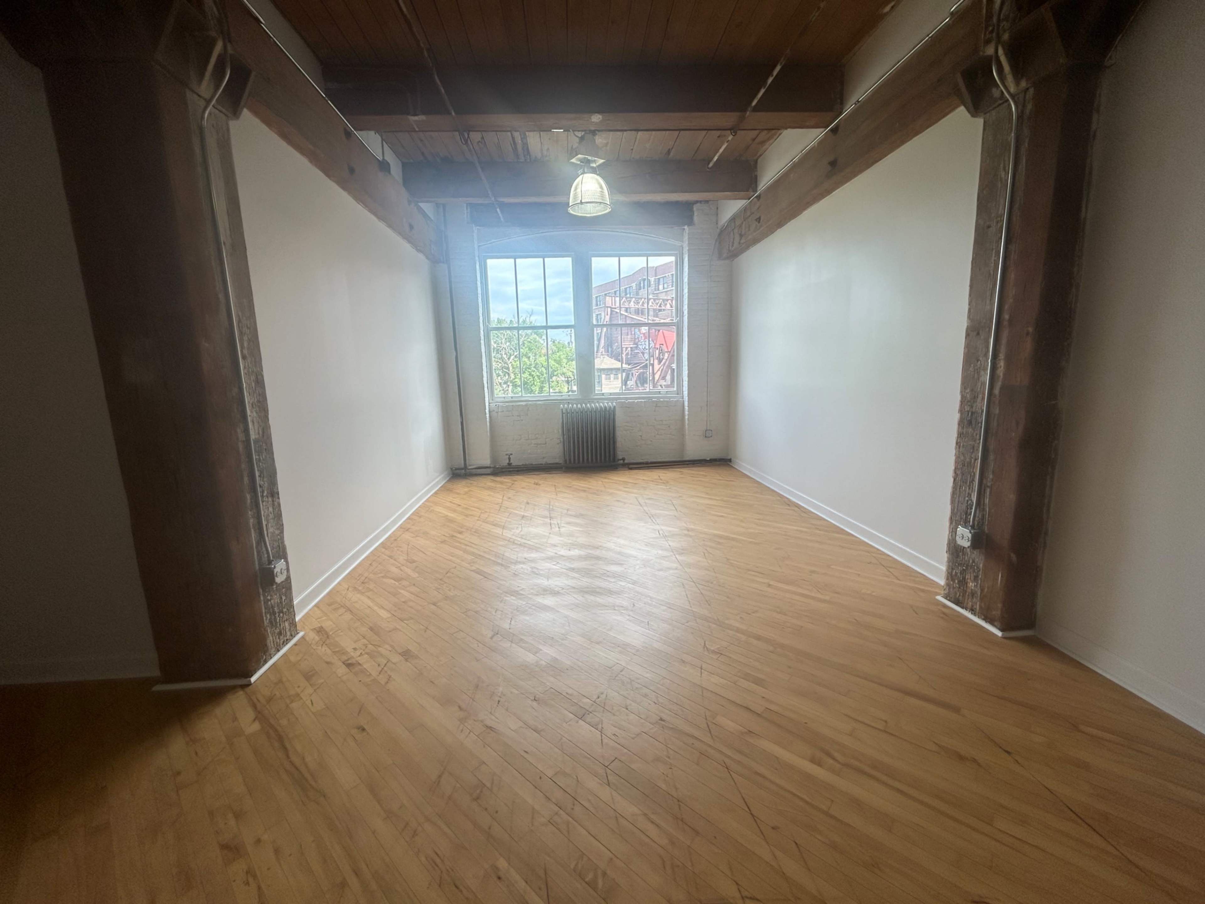 An empty room with wooden floors, exposed beams, and a large window allowing natural light in.