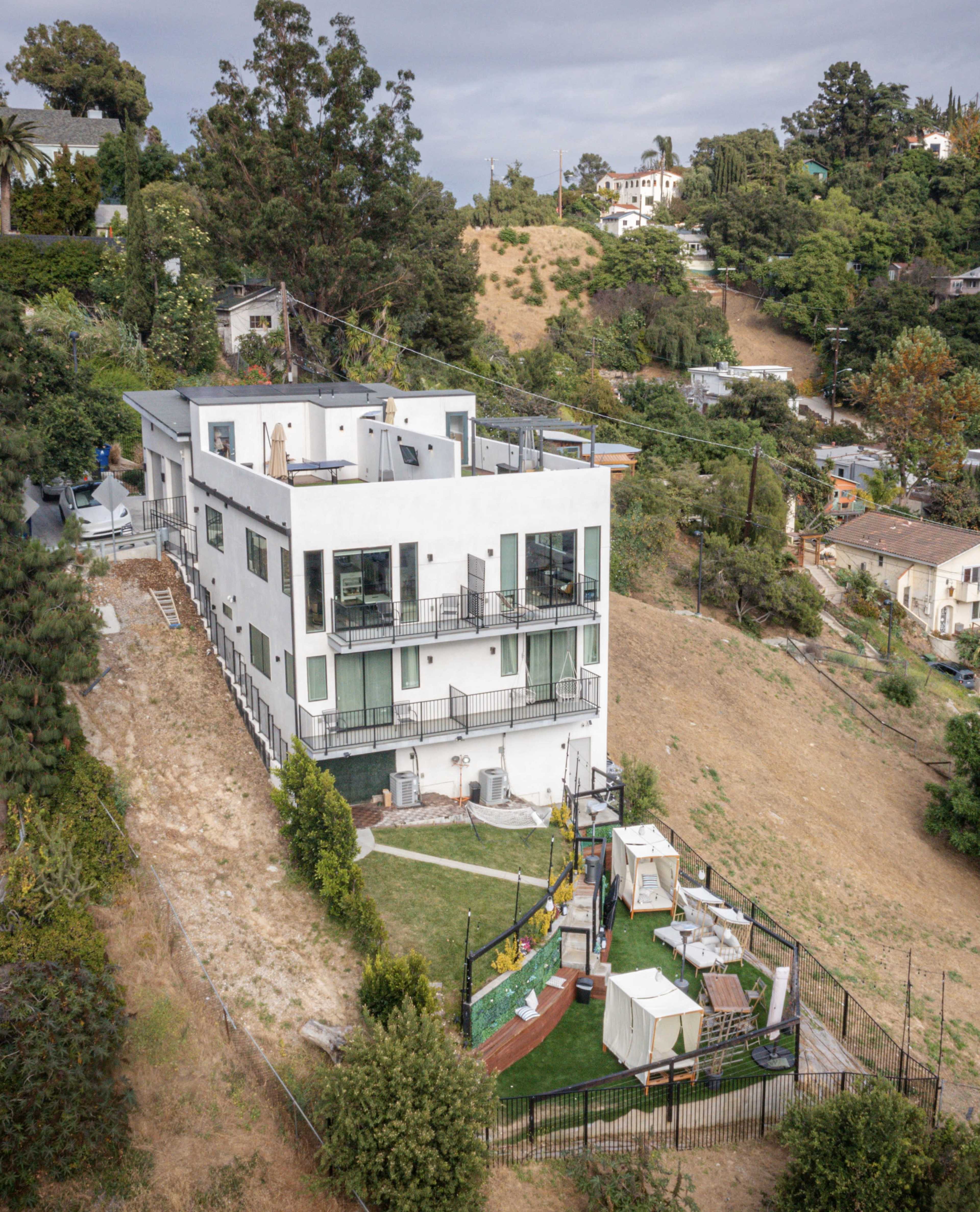 A modern multi-level house with a flat roof overlooks a hillside, featuring a fenced backyard with greenery and outdoor furniture.