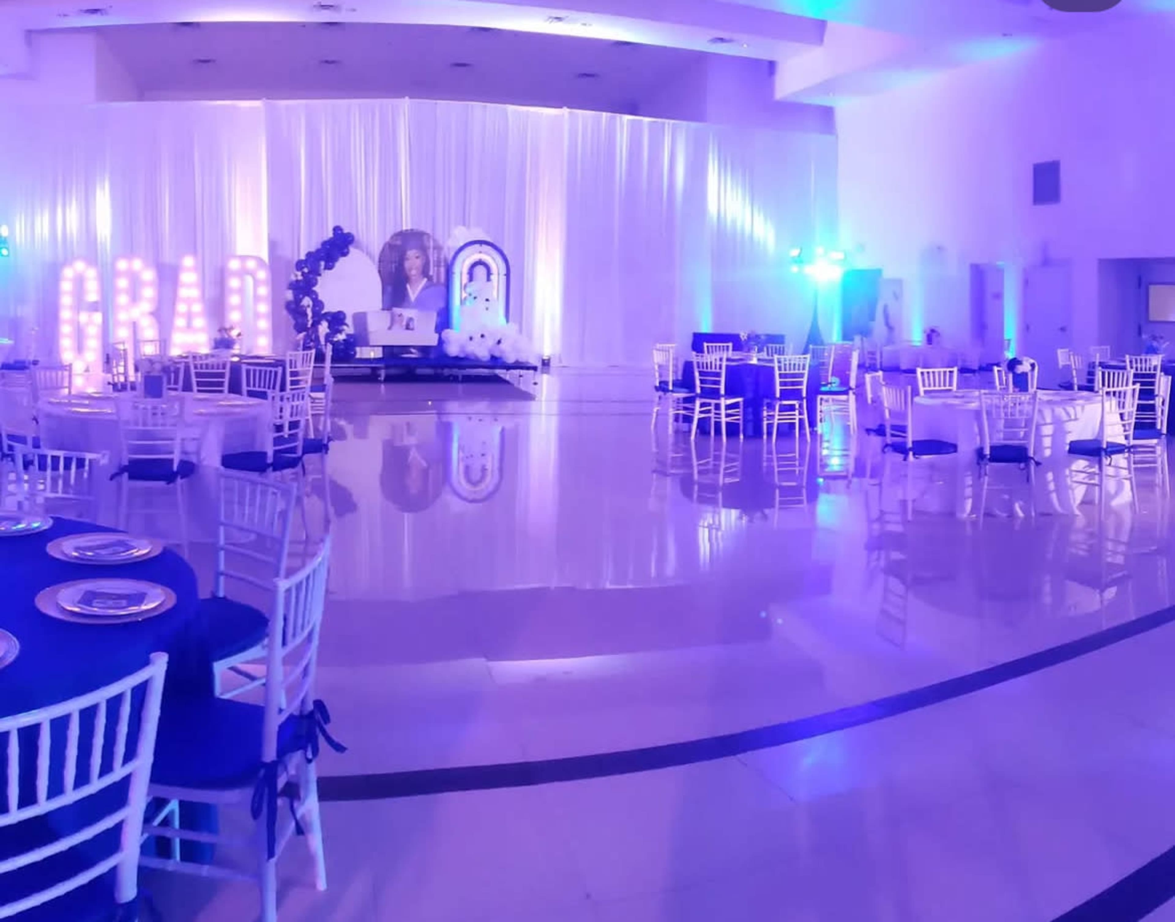 The image shows a decorated events hall with illuminated "GRAD" letters, circular tables with chairs, and a backdrop featuring a large portrait.