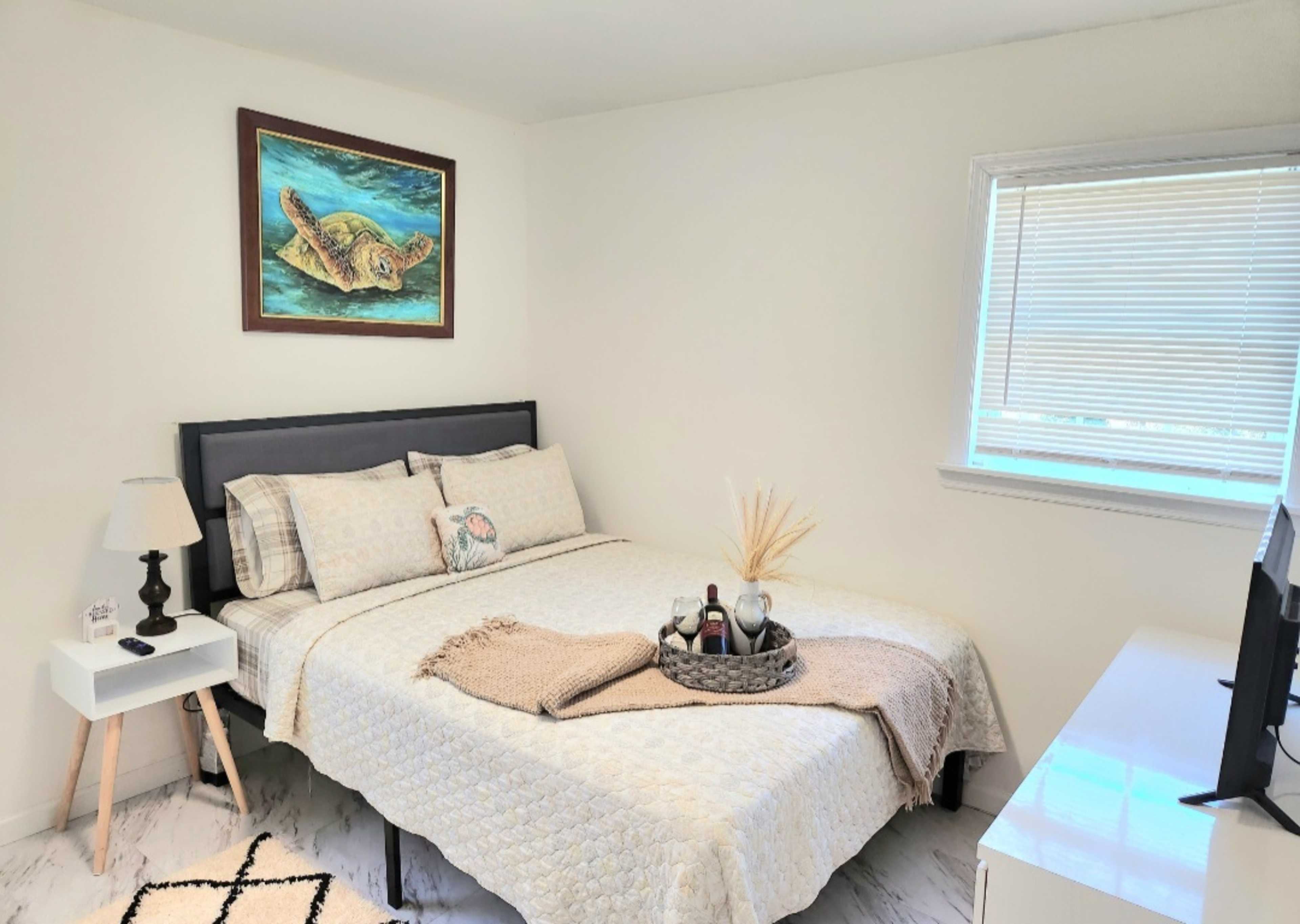 A neatly arranged bedroom featuring a bed with a patterned comforter, a nightstand with a lamp, and a colorful turtle painting above the bed.