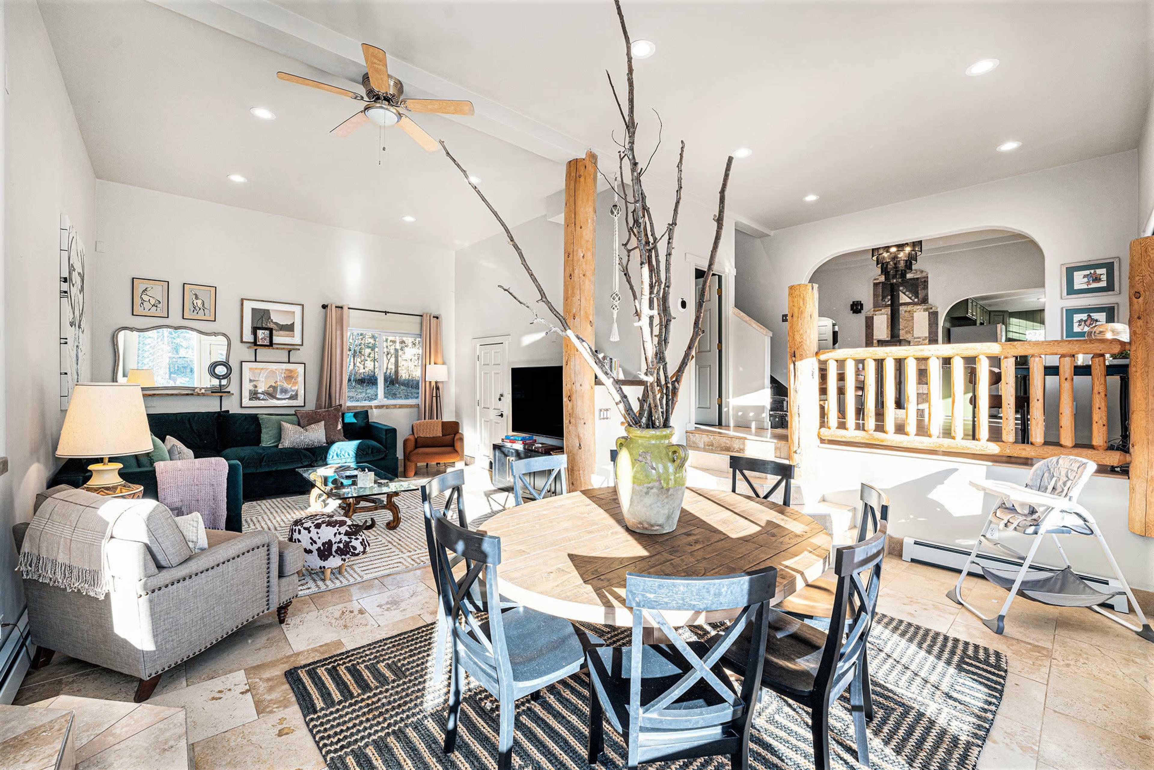 The image shows a modern, open-concept living and dining area with a large round table, a decorative plant center, and a green sofa in a well-lit space featuring wooden beams and a staircase.