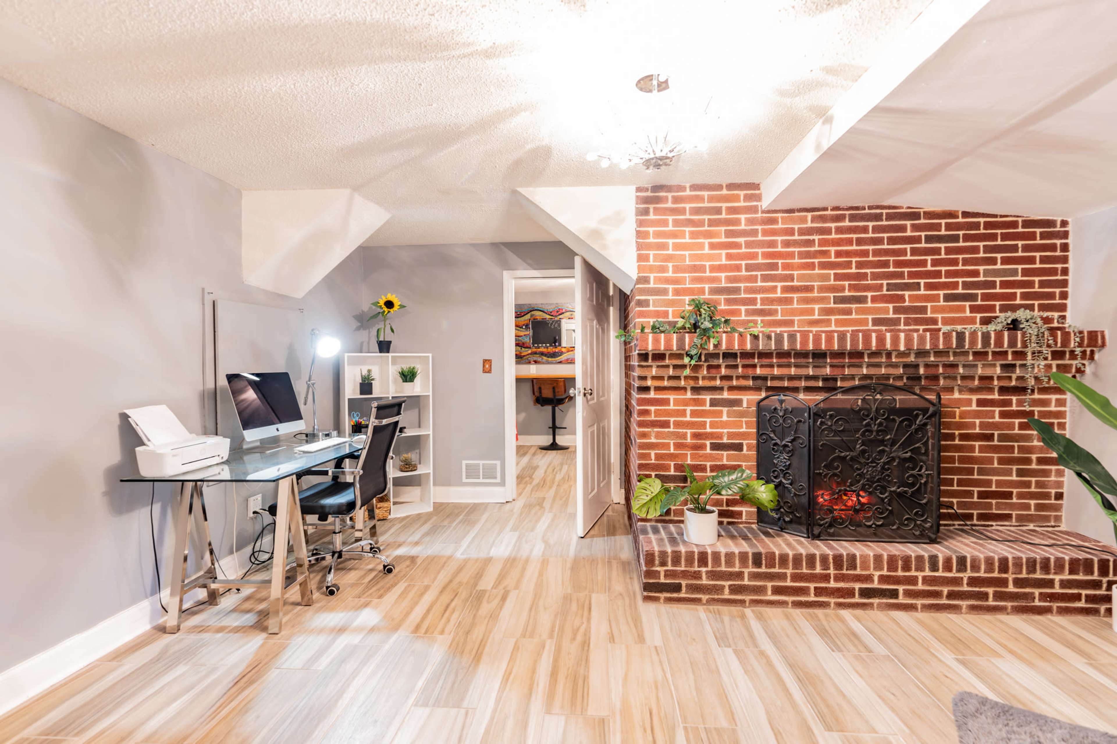The image shows a modern living area featuring a desk with an office chair, a brick fireplace with decorative elements, and a light-colored floor.