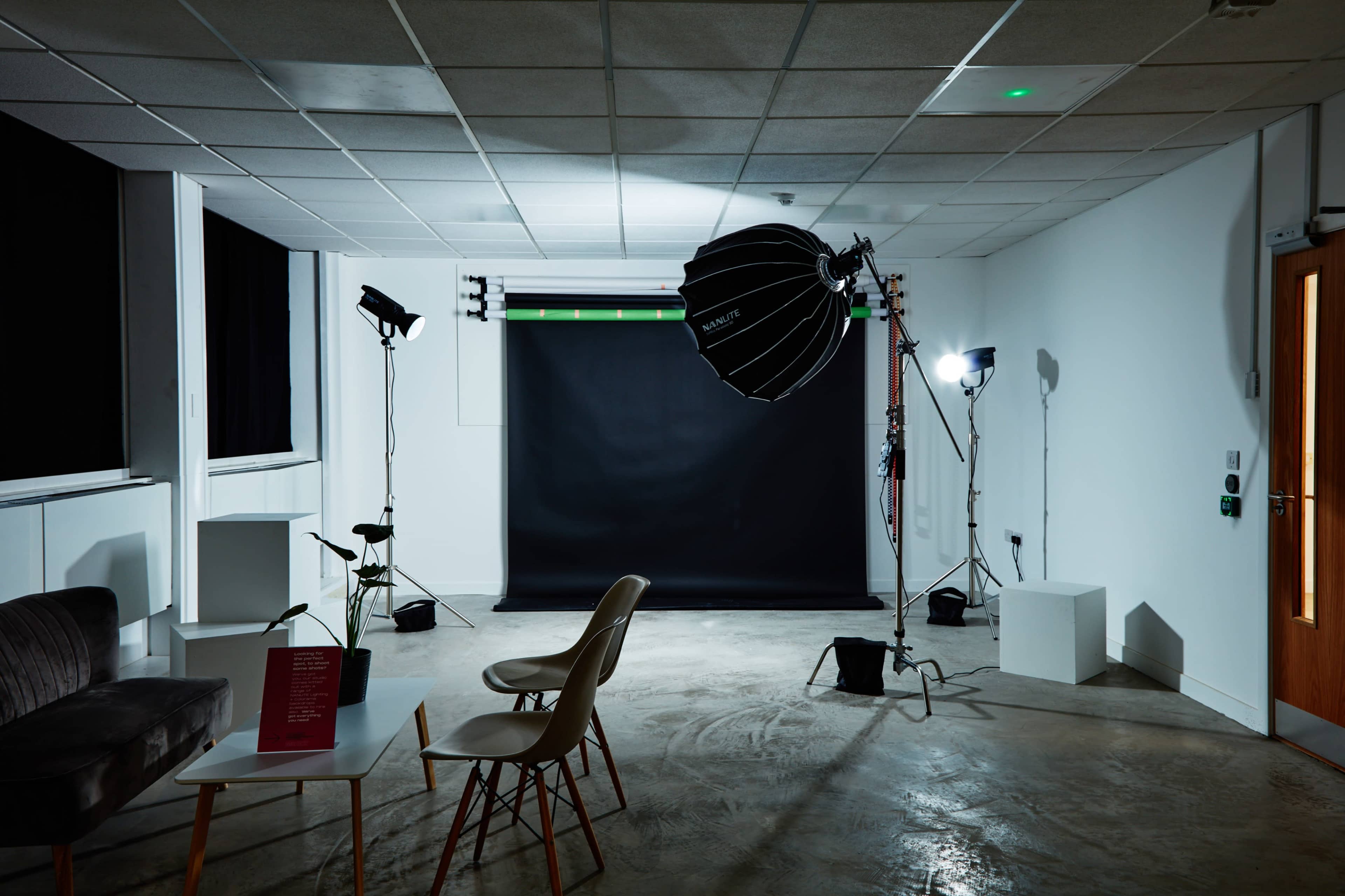 The image shows a photography studio with a black backdrop, lighting equipment, and a few pieces of furniture including a sofa and chairs.