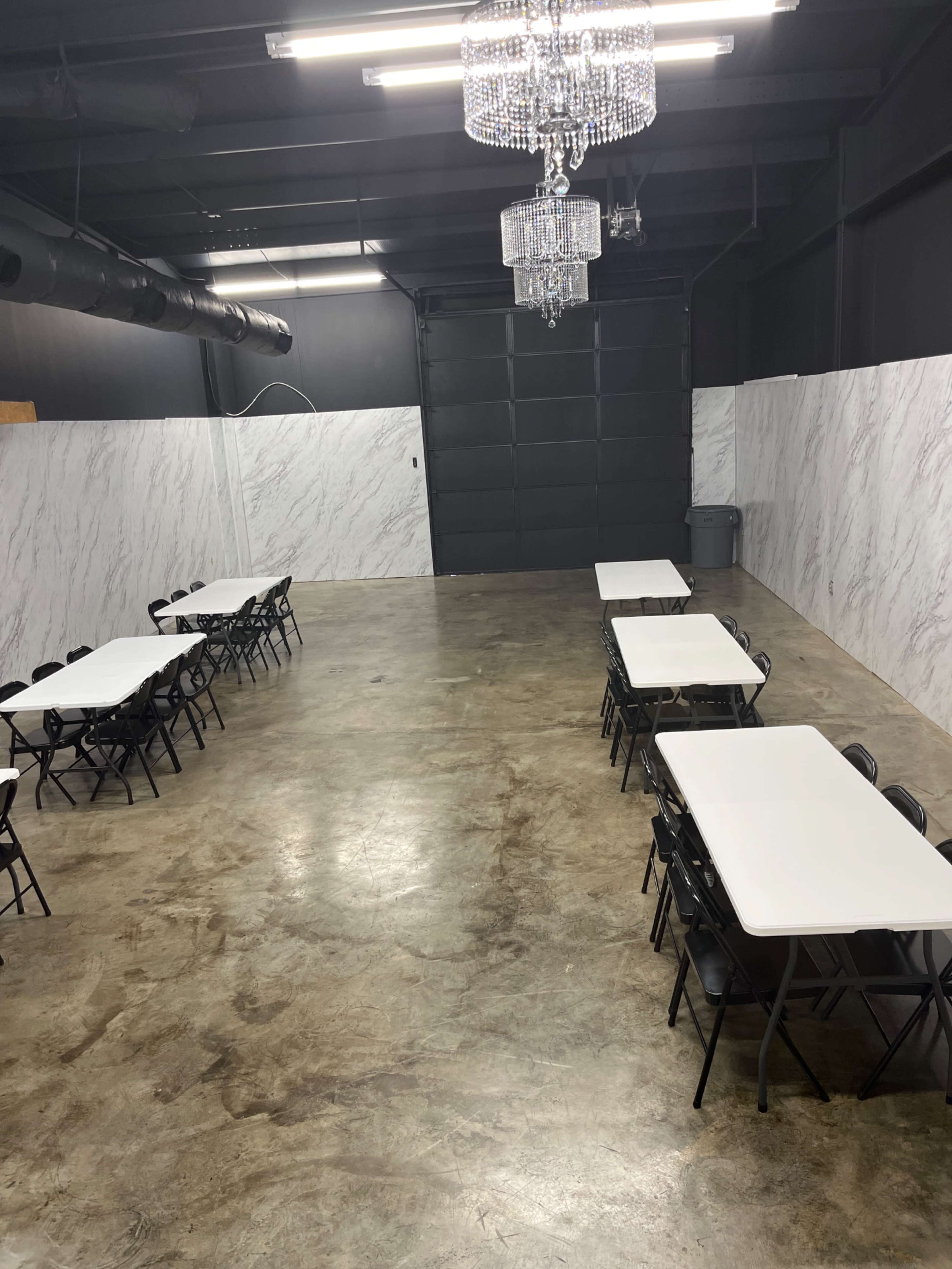A spacious room with polished concrete floors, featuring several rows of white tables and black chairs, and two chandeliers hanging from the ceiling.