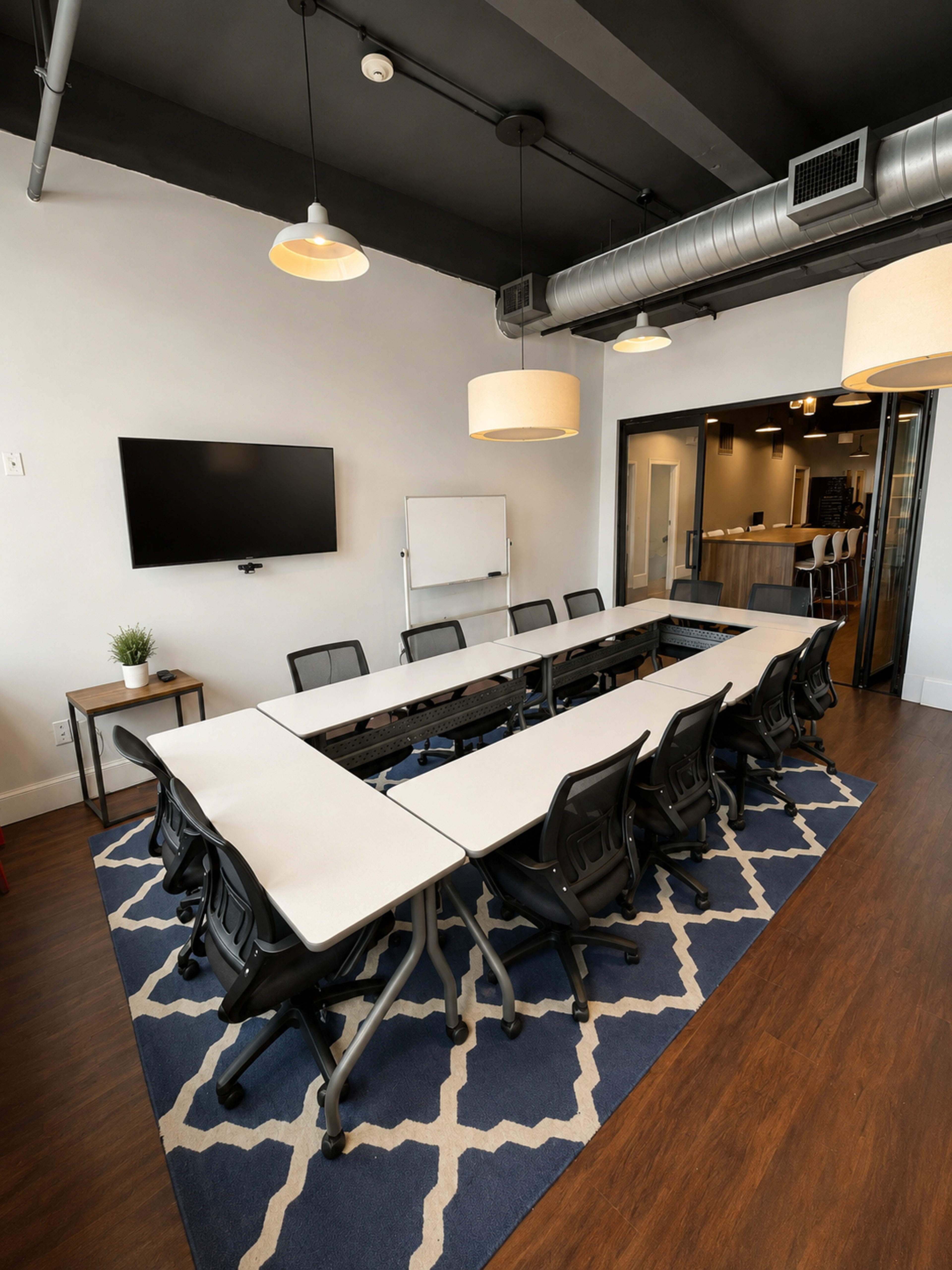 The image shows a modern conference room with a long table surrounded by chairs, a large TV mounted on the wall, and a whiteboard on a stand.