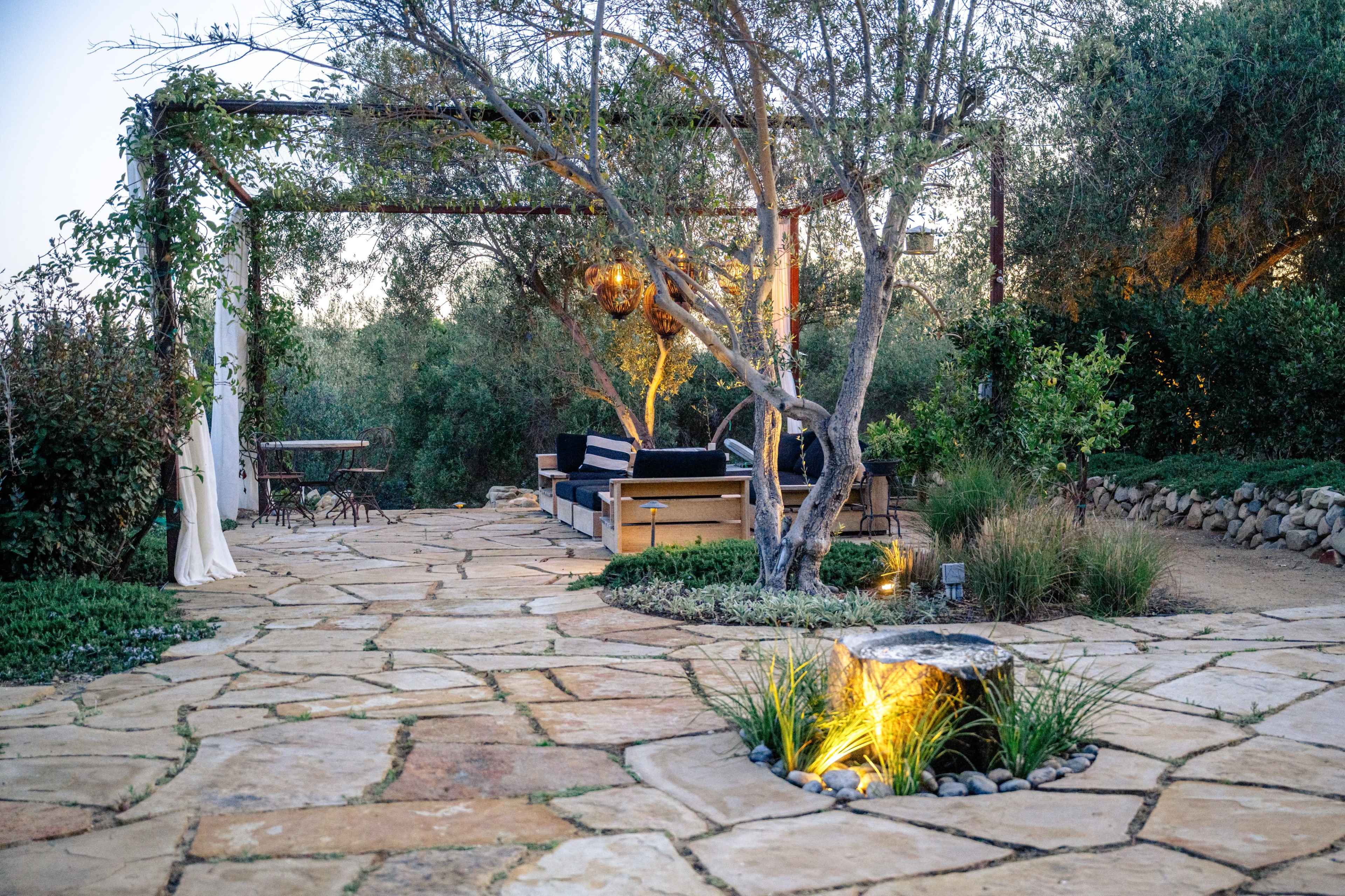 The image shows a landscaped outdoor area featuring a stone pathway, a seating arrangement under a pergola, and surrounding greenery.