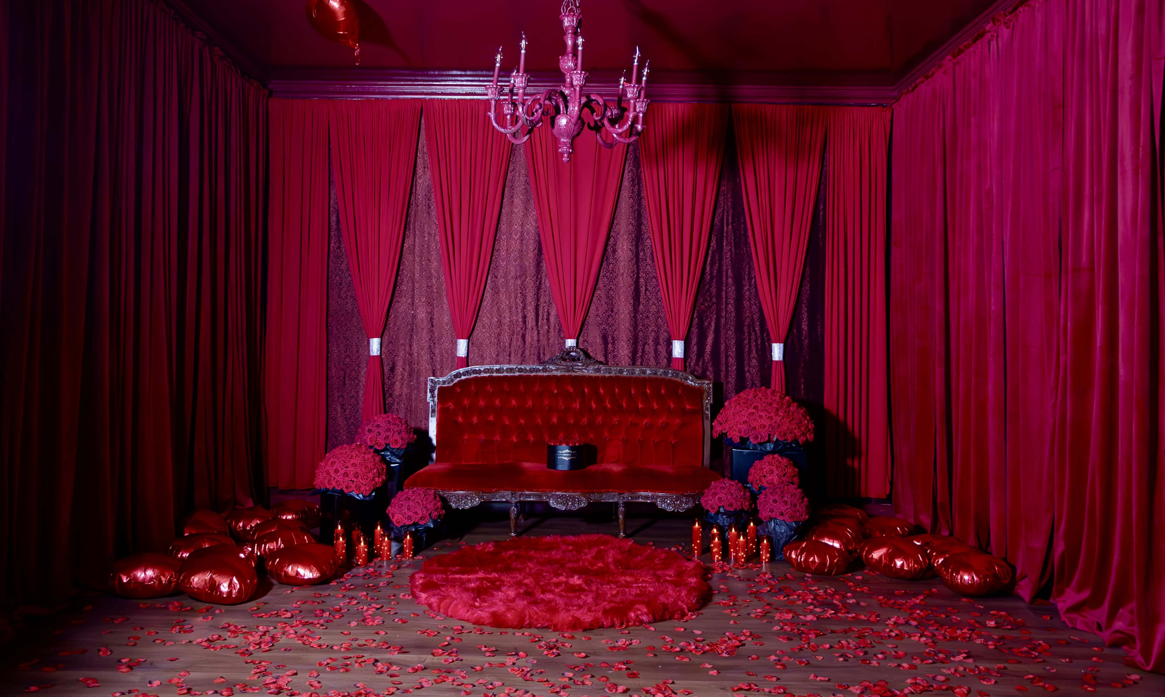 The scene features a richly decorated room with red walls, a velvet sofa, and an array of red decorations, including flowers and pillows scattered across the floor.