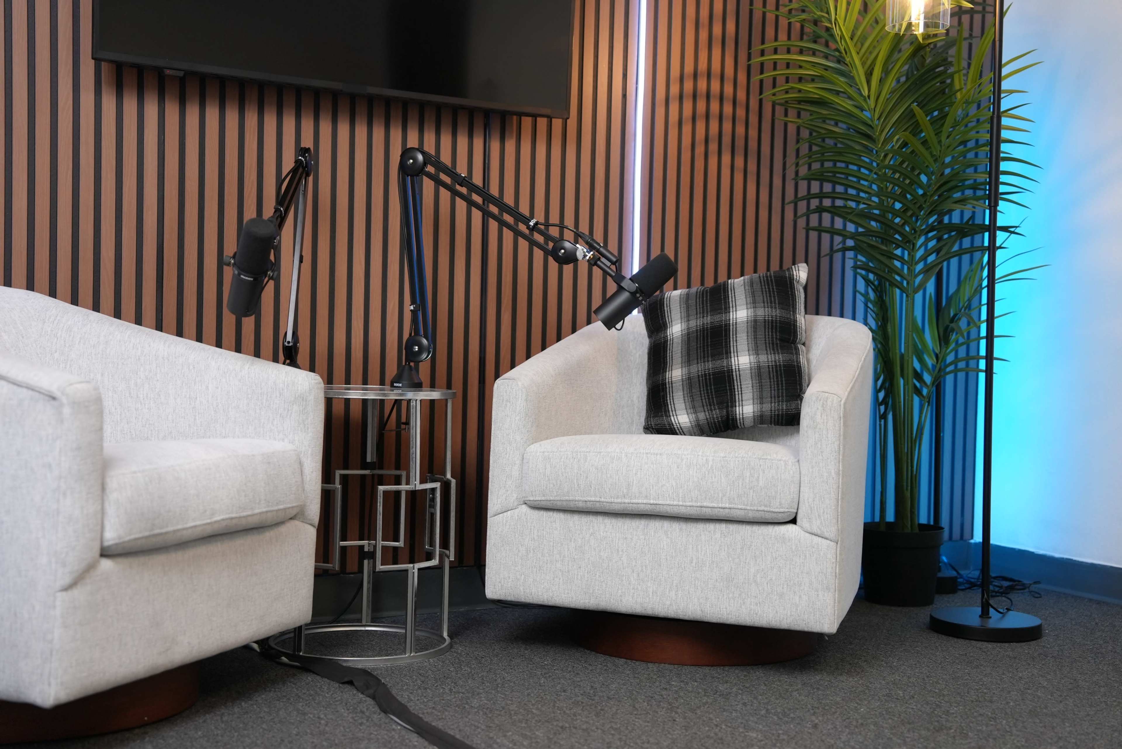 The image features two modern, light gray swivel chairs positioned next to a small table, with a plant and lighted backdrop in a studio setting.