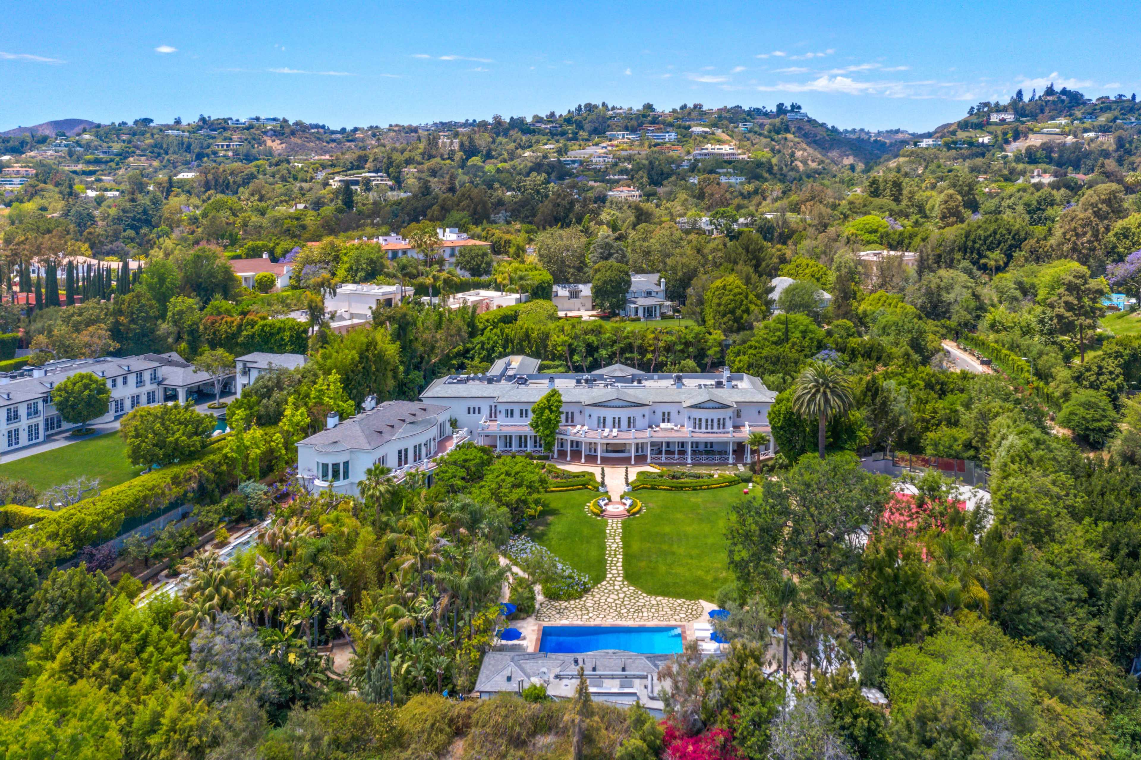 A large, luxurious mansion with a sprawling lawn and swimming pool, surrounded by lush greenery and hills in the background.