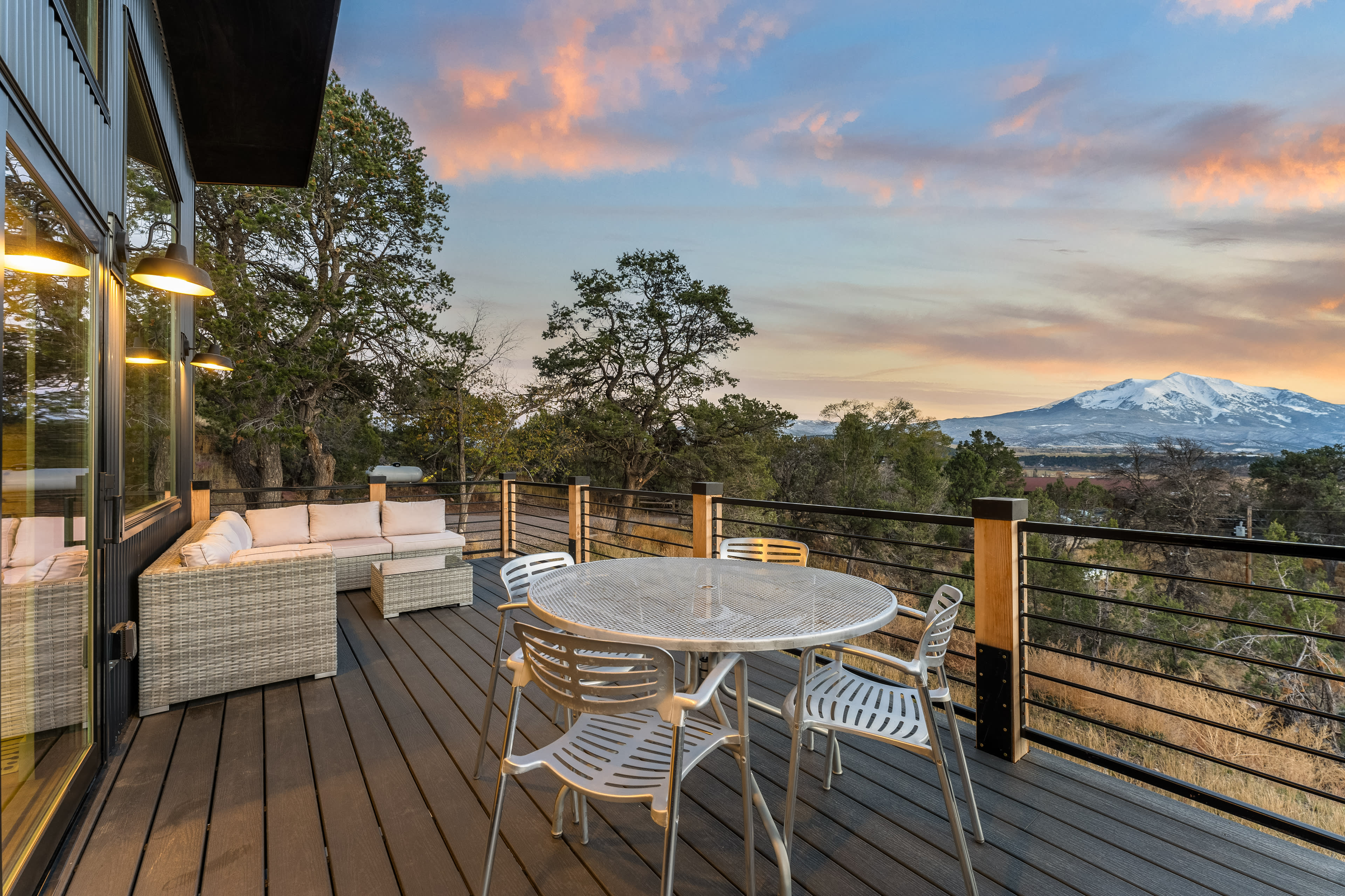 The image shows a wooden deck with a round table and chairs, overlooking a mountain landscape at sunset.