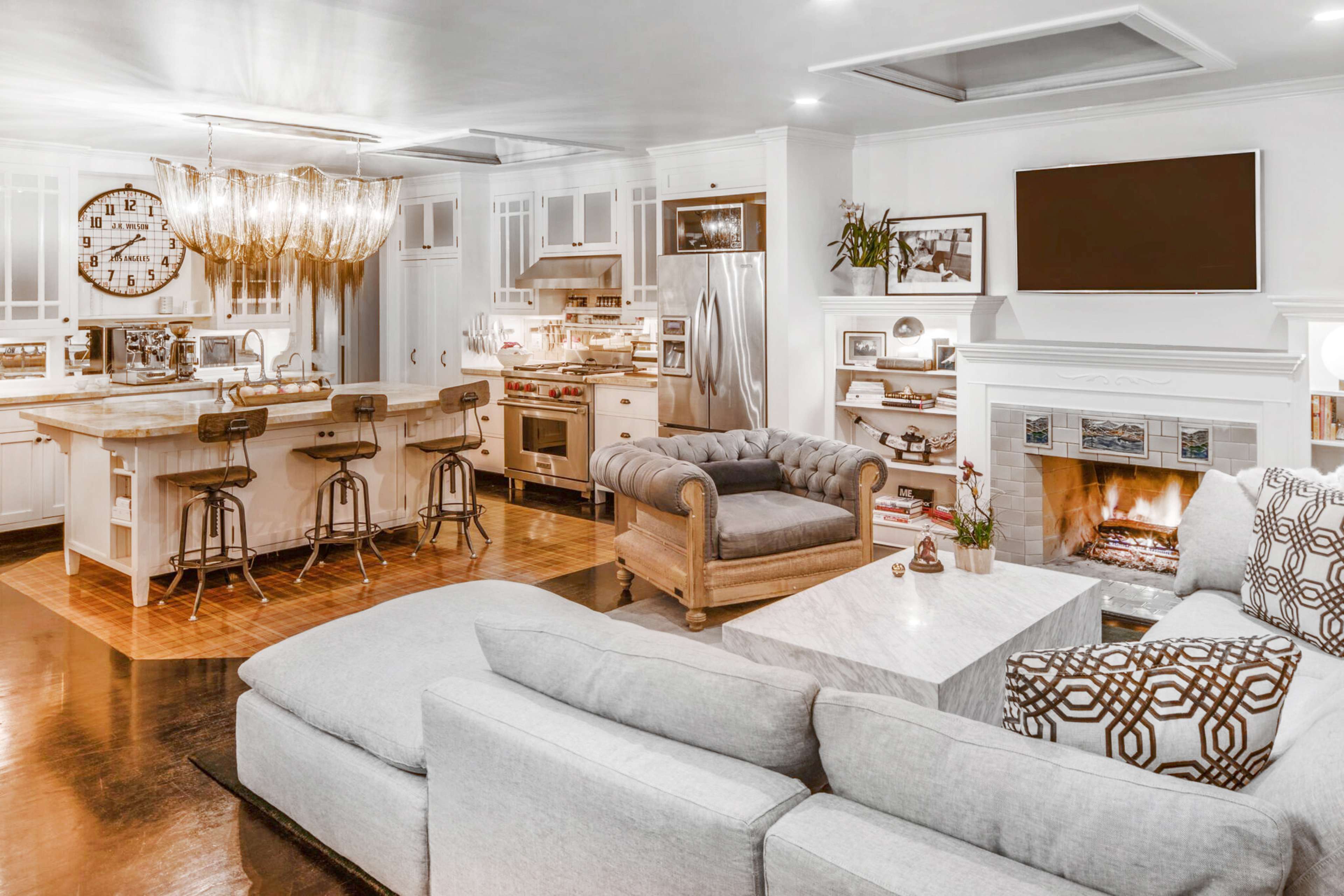 The image shows a modern living area with a gray sectional sofa, a marble coffee table, and an open kitchen featuring a large island with bar stools and stainless steel appliances.