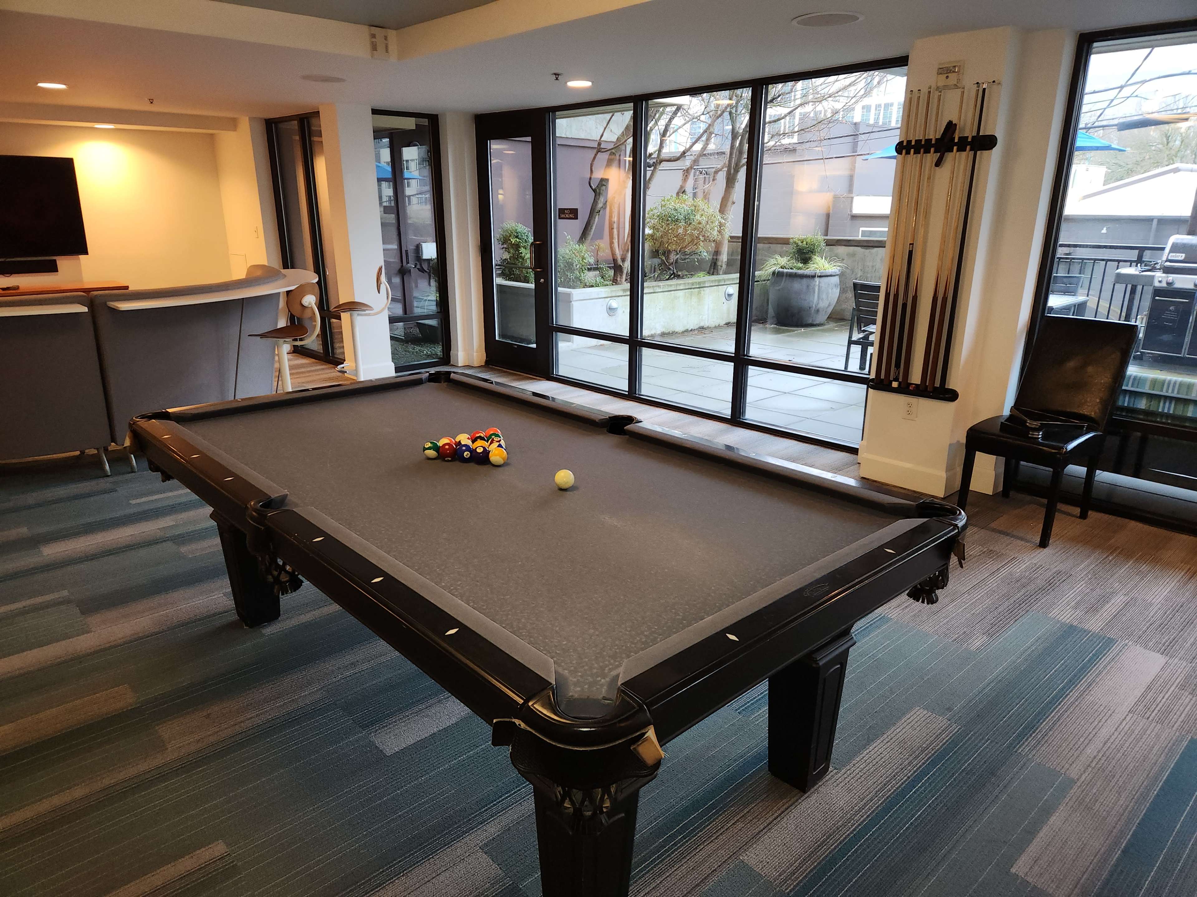 A billiards table is centered in a room with large windows overlooking an outdoor patio area.