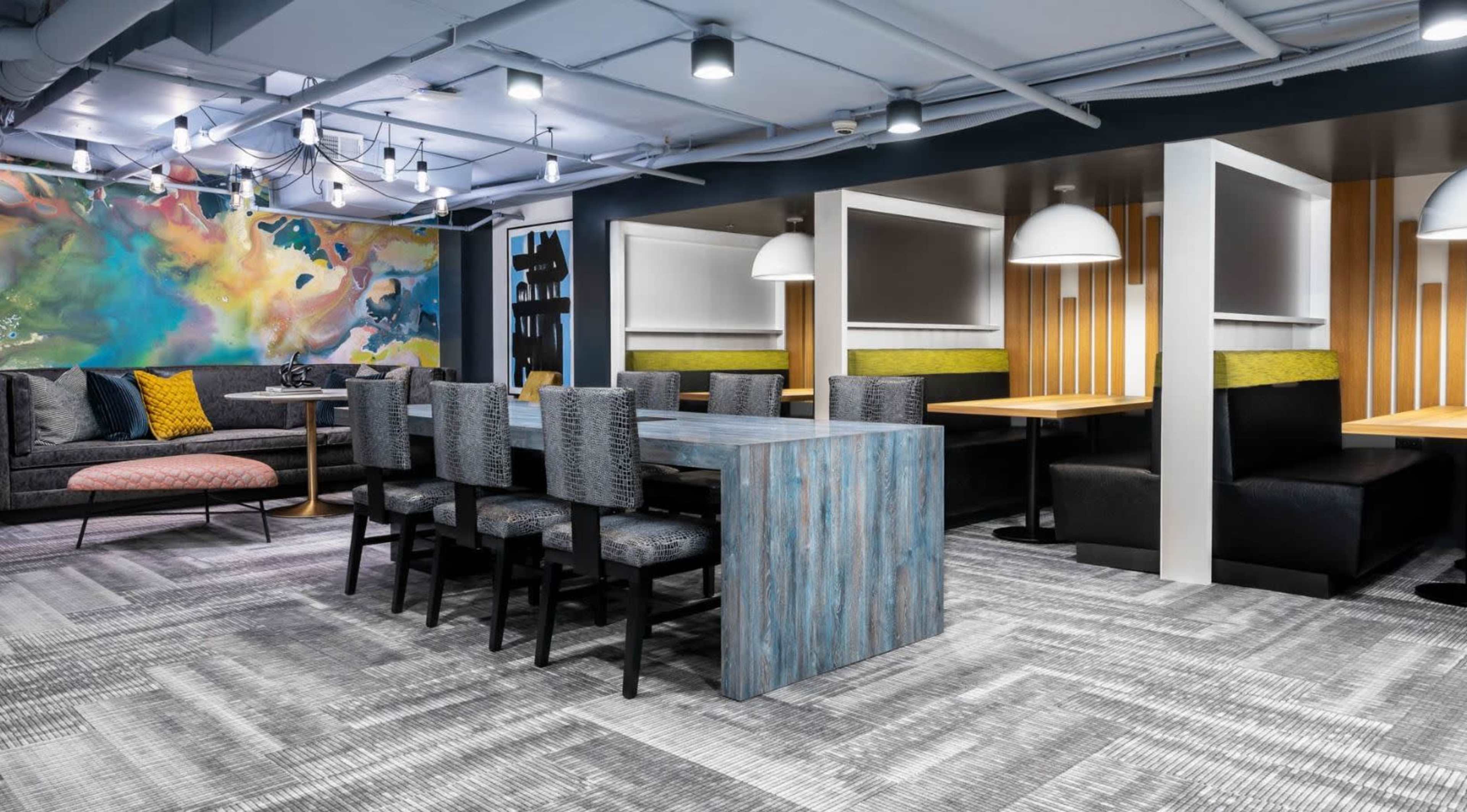 A modern coworking space featuring a long table with black chairs, colorful wall art, and private booth seating areas.