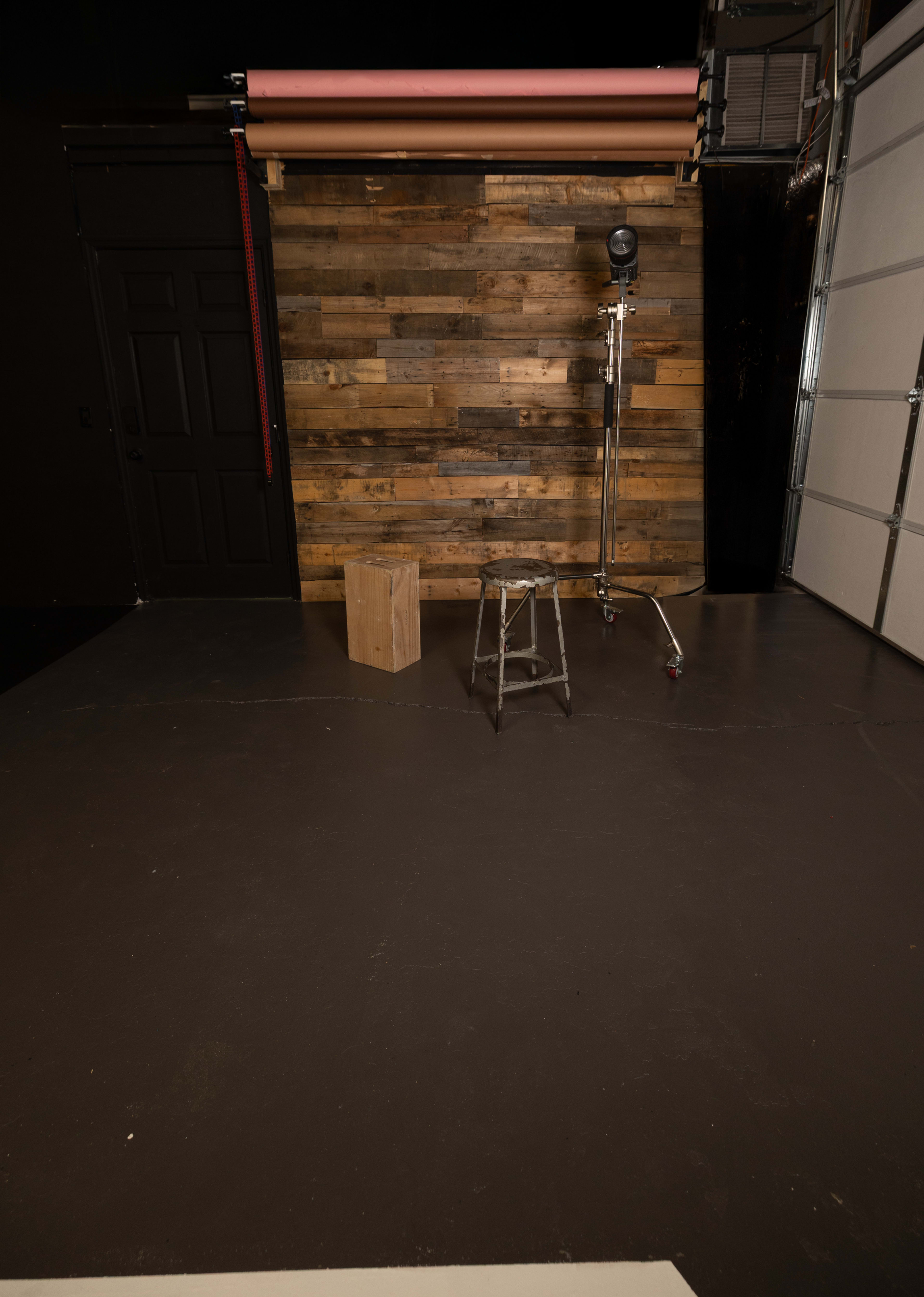 The image depicts a photography studio with a wooden backdrop, a spotlight on a stand, a metal ladder, and a wooden box on the floor.
