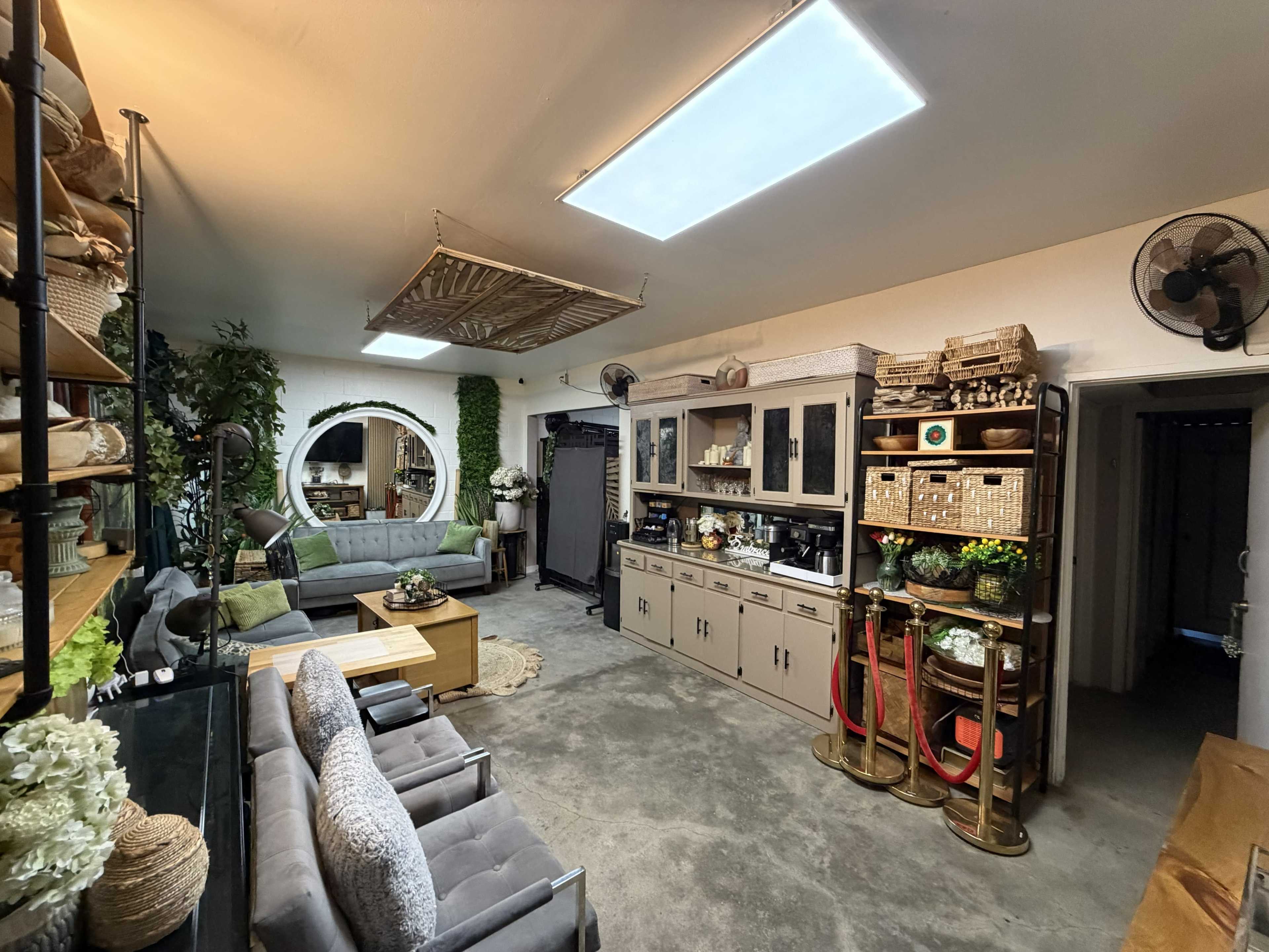 The image shows a modern interior space designed as a cozy living area and workspace, featuring comfortable seating, decorative plants, and organized shelves with various items.