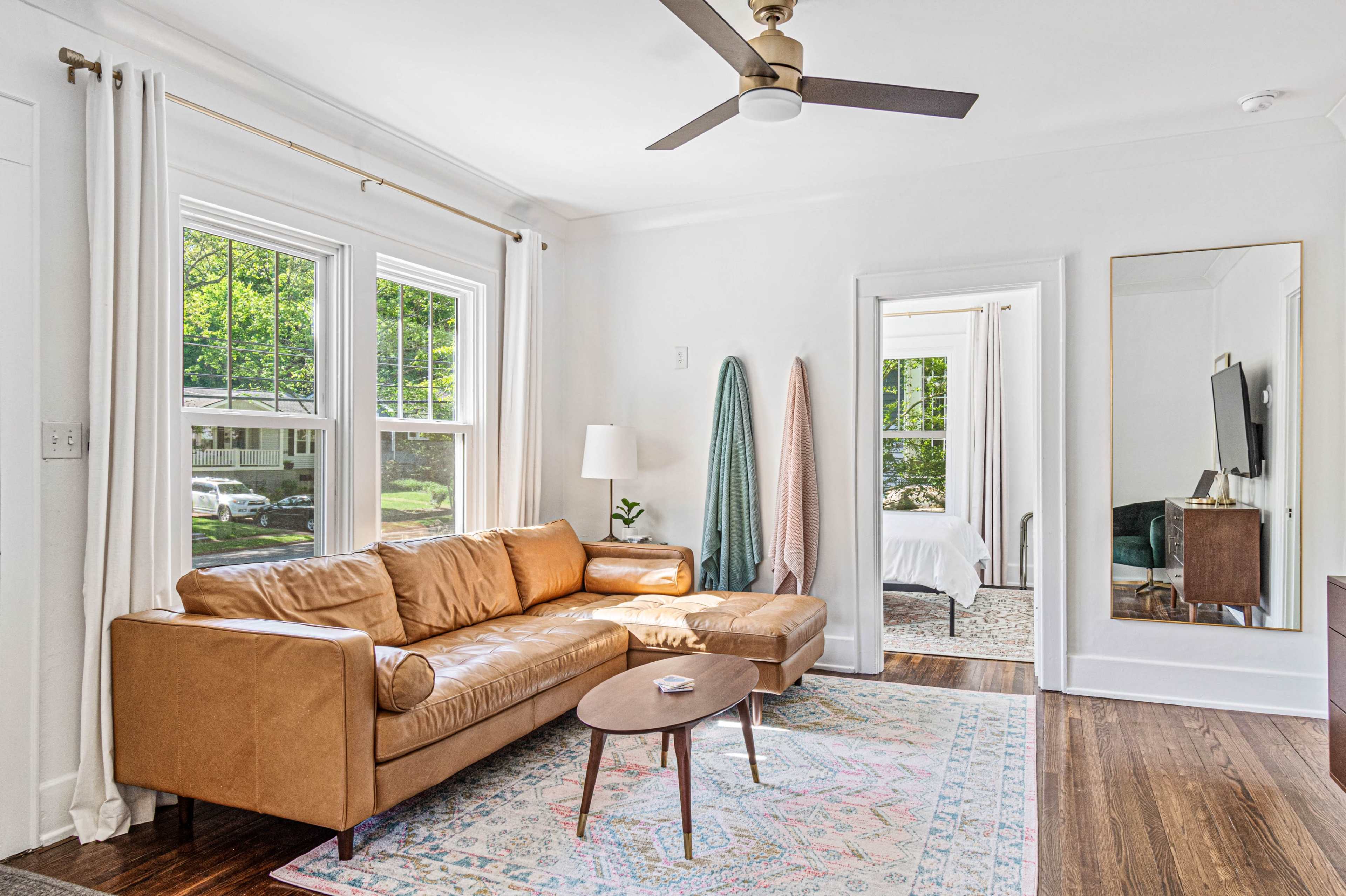 The living room features a tan leather sectional couch, a small round coffee table, and a large mirror, with sunlight streaming through the windows.