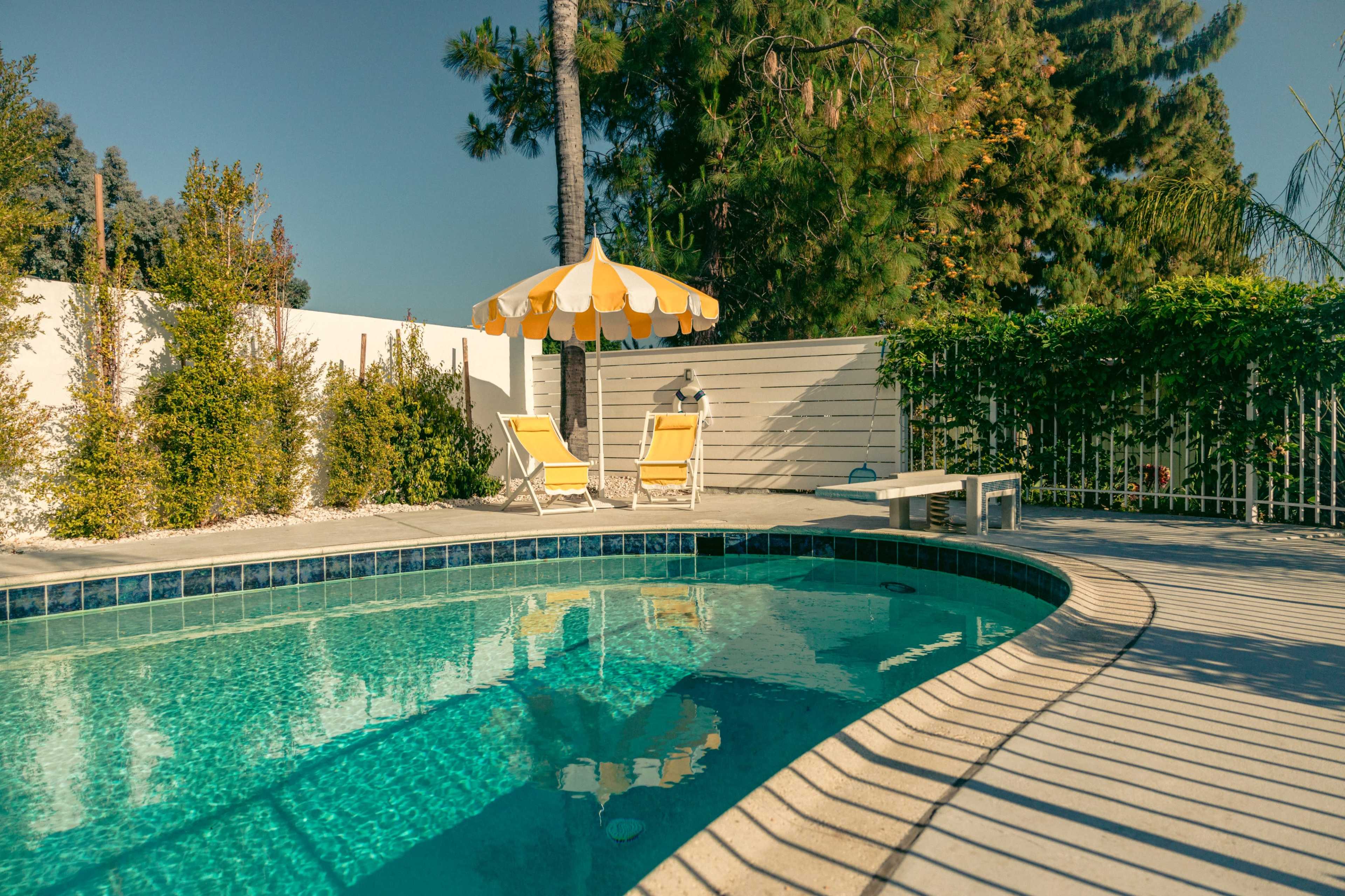 A clear swimming pool is surrounded by lounge chairs and a sun umbrella, with greenery and a white fence in the background.