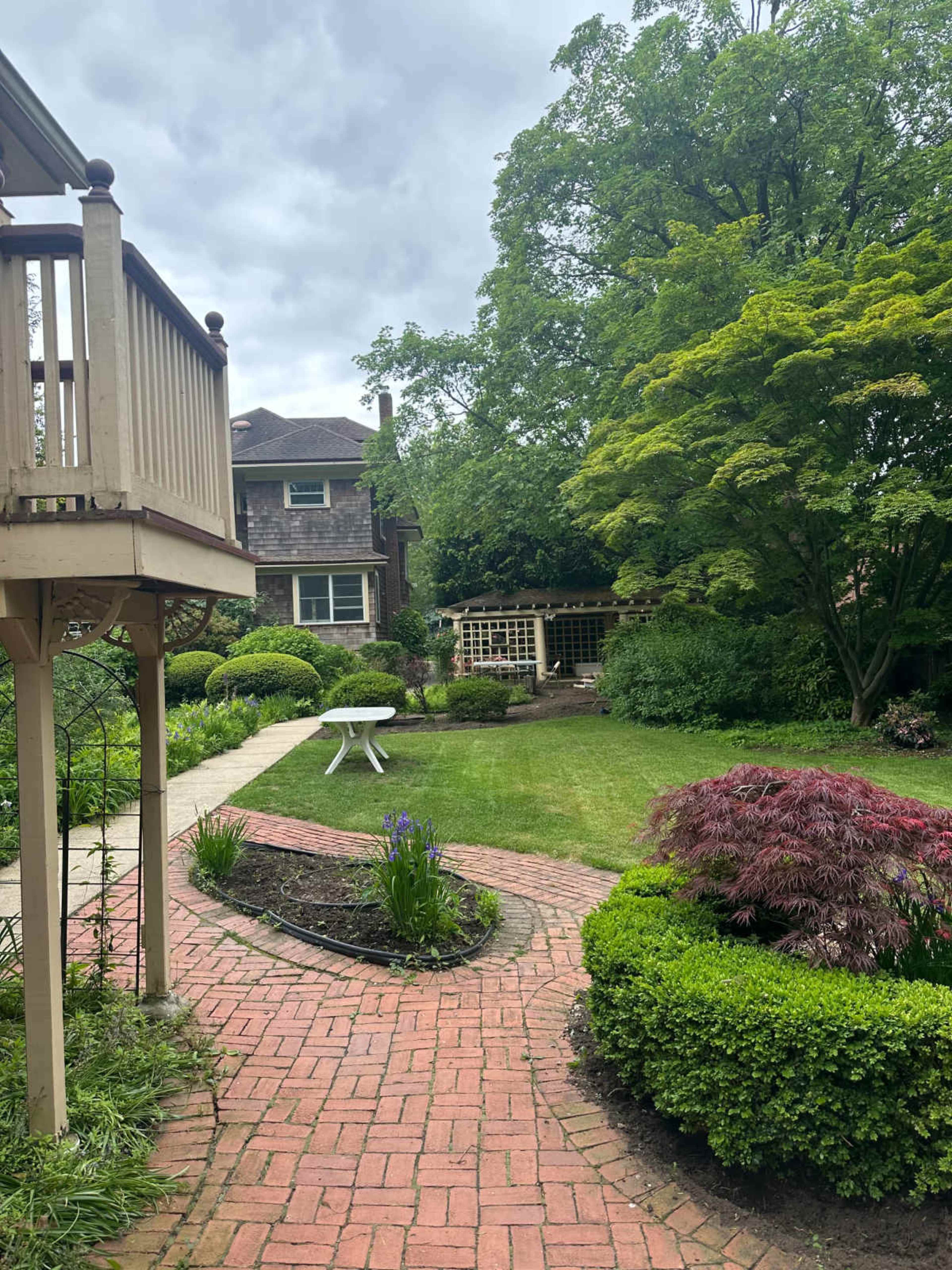 A garden path made of brick leads to a green lawn area surrounded by various shrubs and trees, with a small white table in the center.