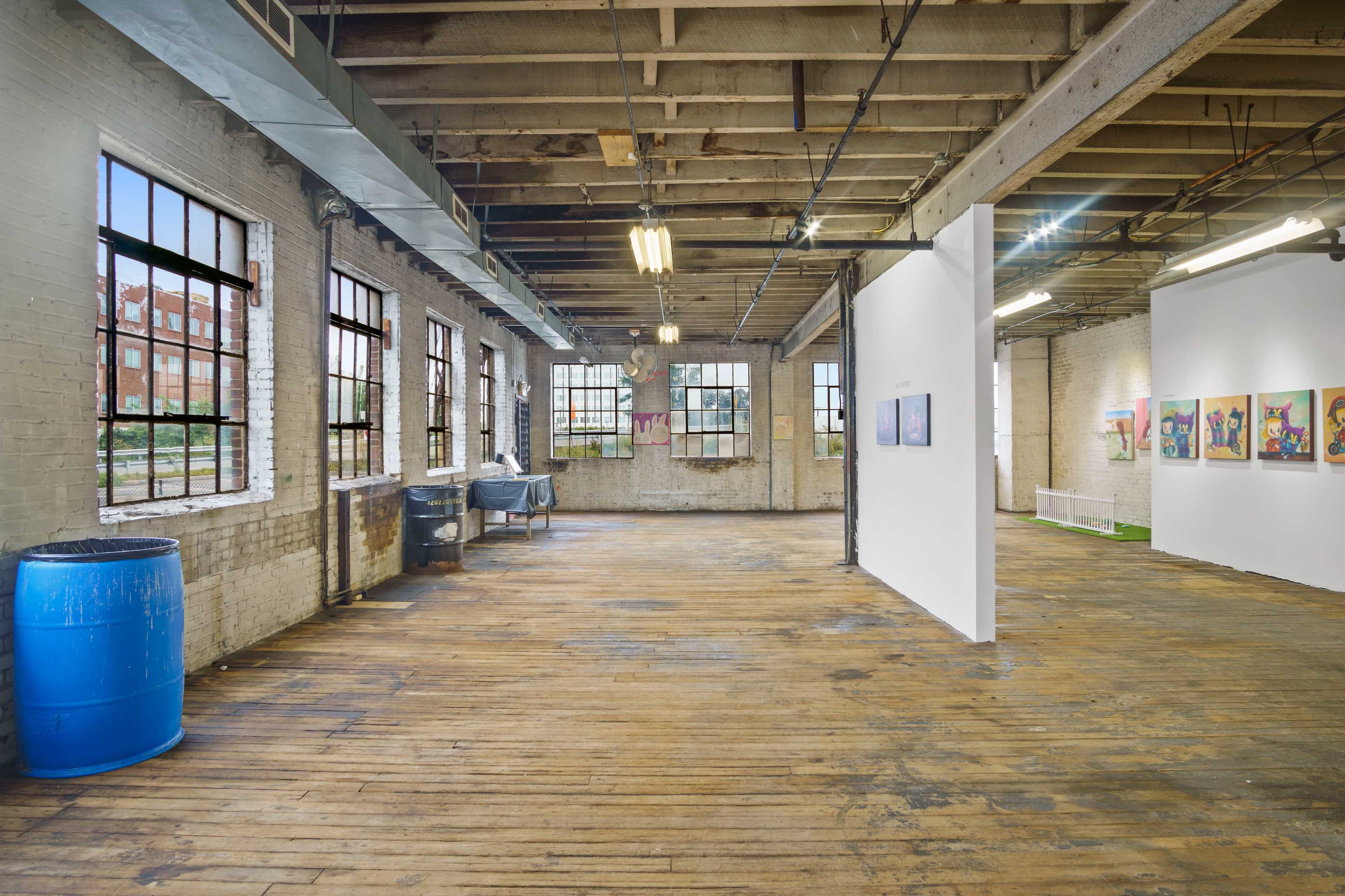 An open art gallery space with wooden floors, exposed brick walls, large windows, and a central partition displaying artwork.
