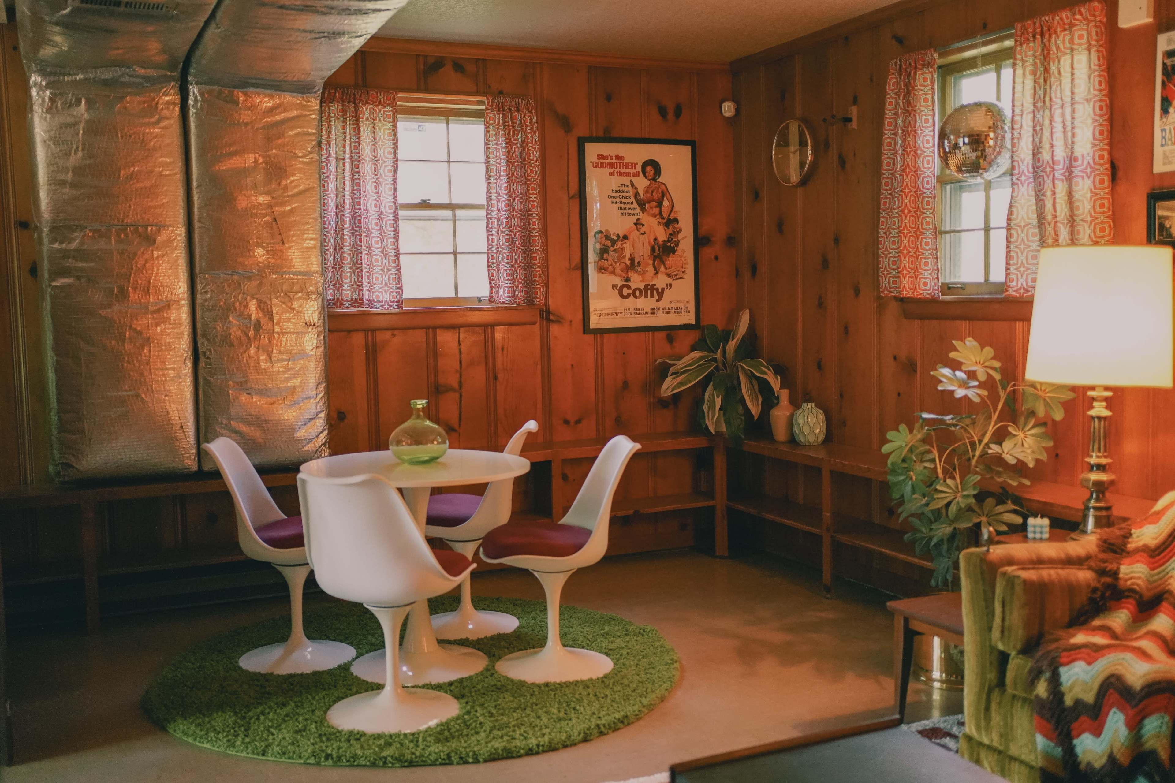A cozy, paneled room with a round table and four white chairs on a green rug, surrounded by decorative plants and vintage posters.