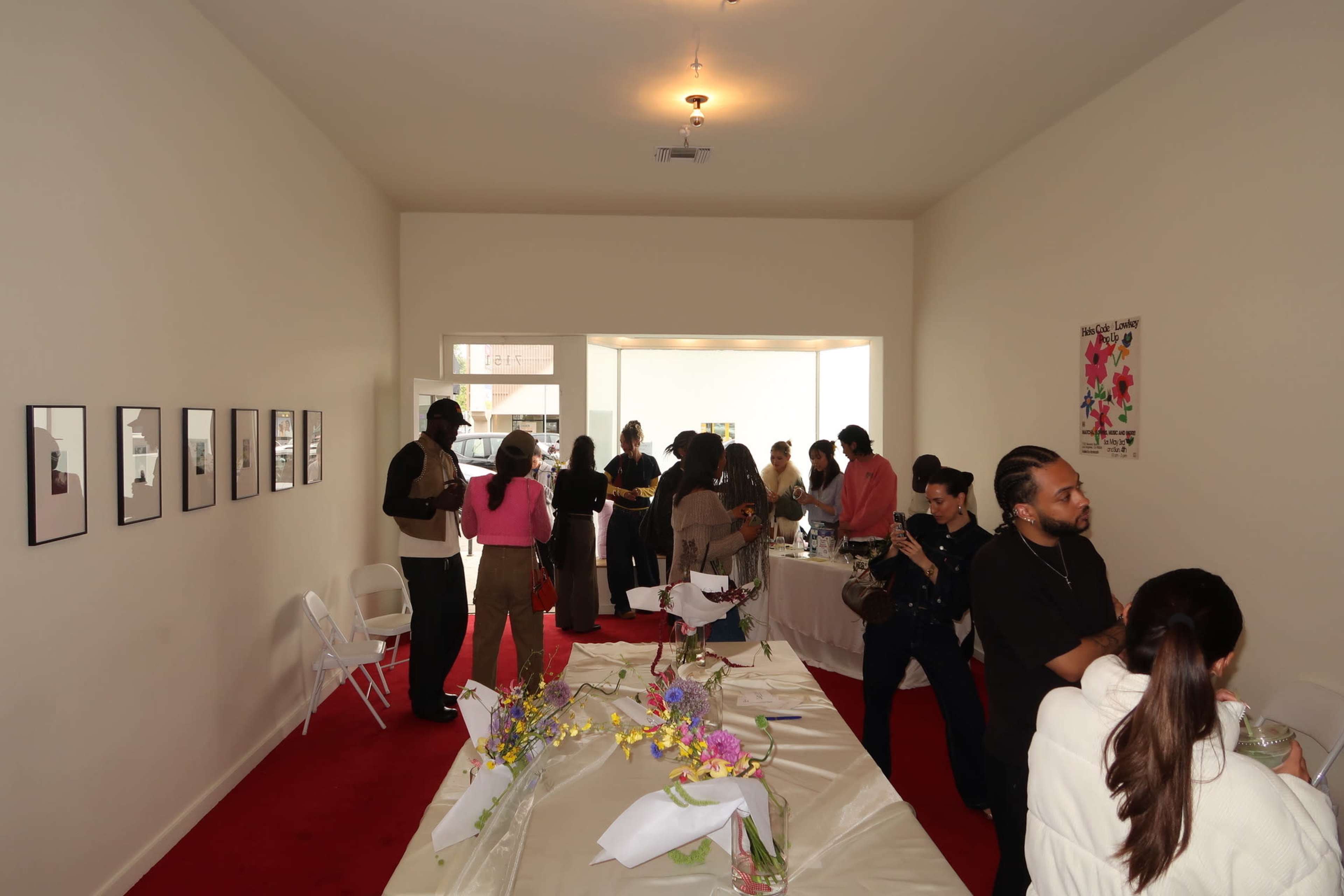 The image shows a gallery-like space with people mingling around tables decorated with floral arrangements and photographs displayed on the walls.