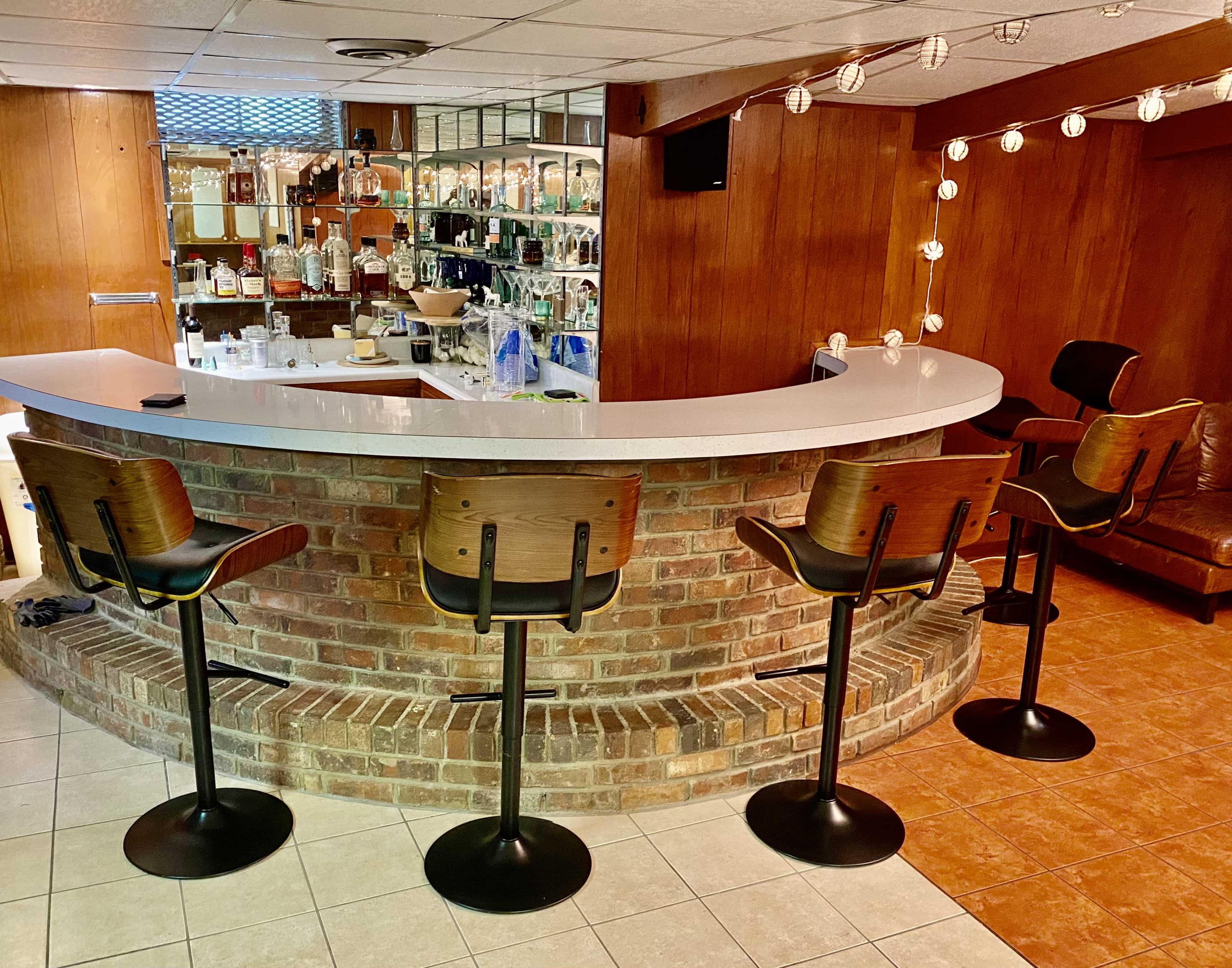 A spacious home bar area with a curved countertop made of white stone, surrounded by four tall wooden stools on a brick base, and a glass shelf displaying various bottles in the background.