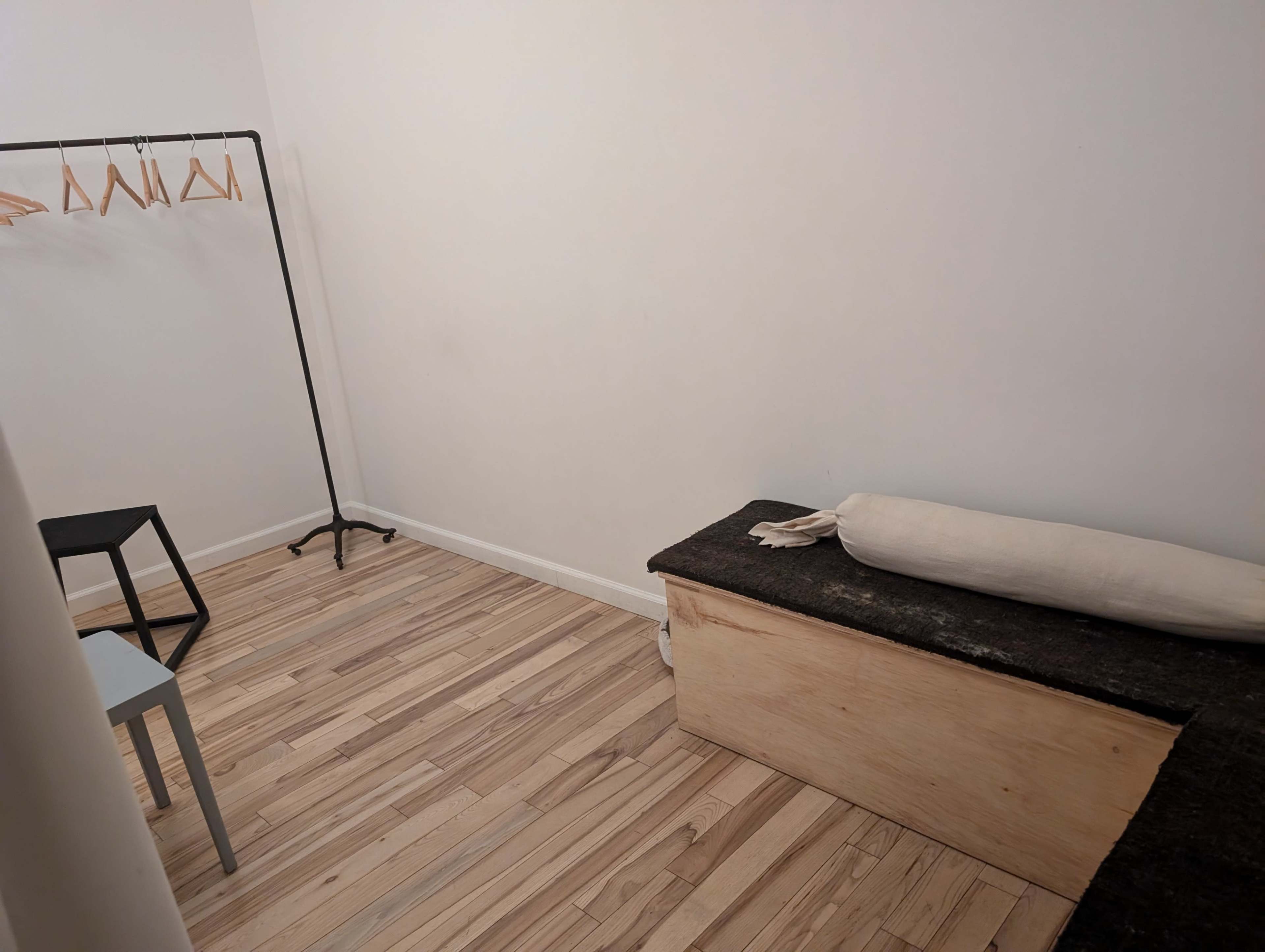 The image shows a minimalistic room with a wooden floor, featuring a clothing rack, a chair, and a storage bench.