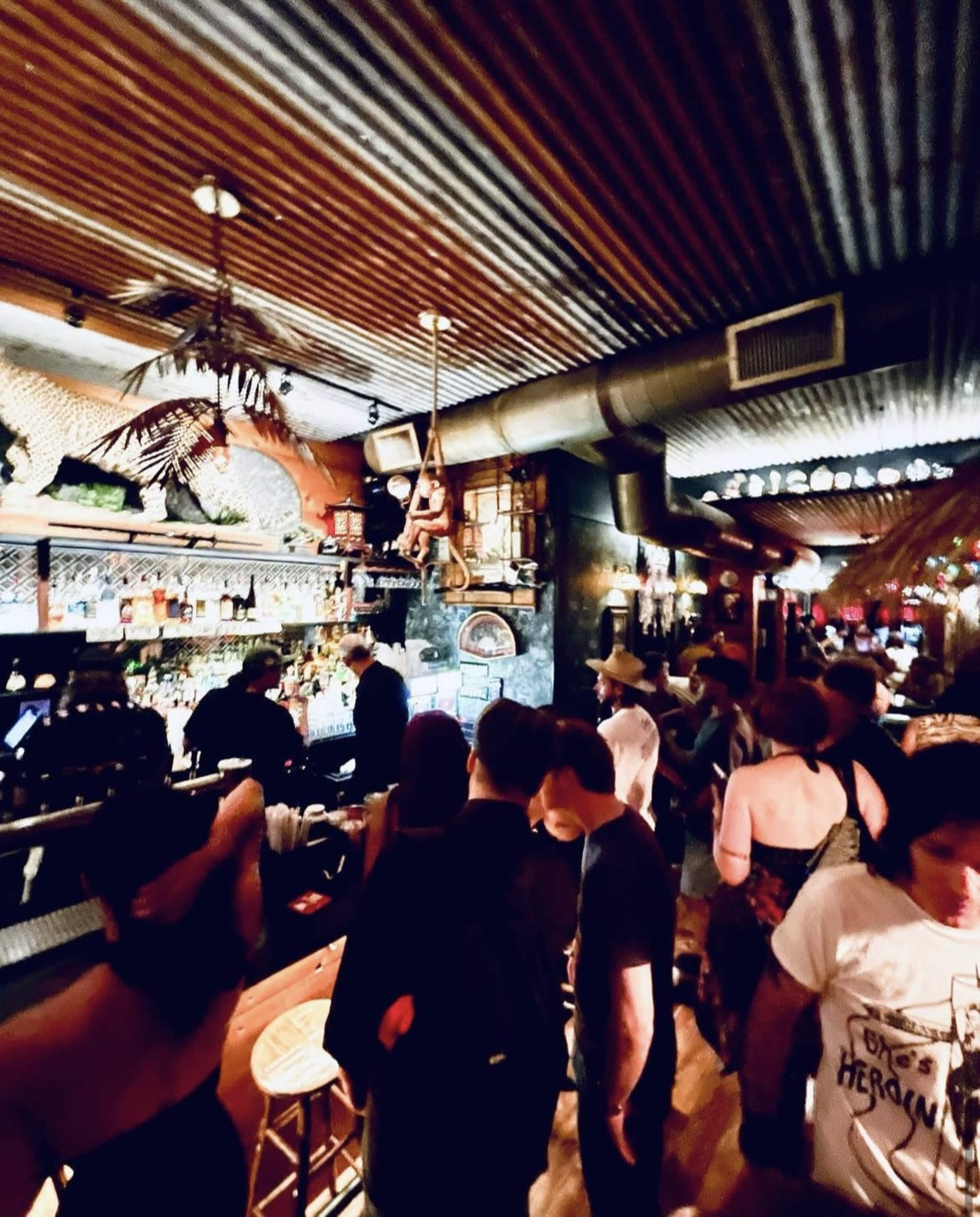 A crowded bar scene shows patrons waiting at the counter while staff prepare drinks in a dimly lit atmosphere.