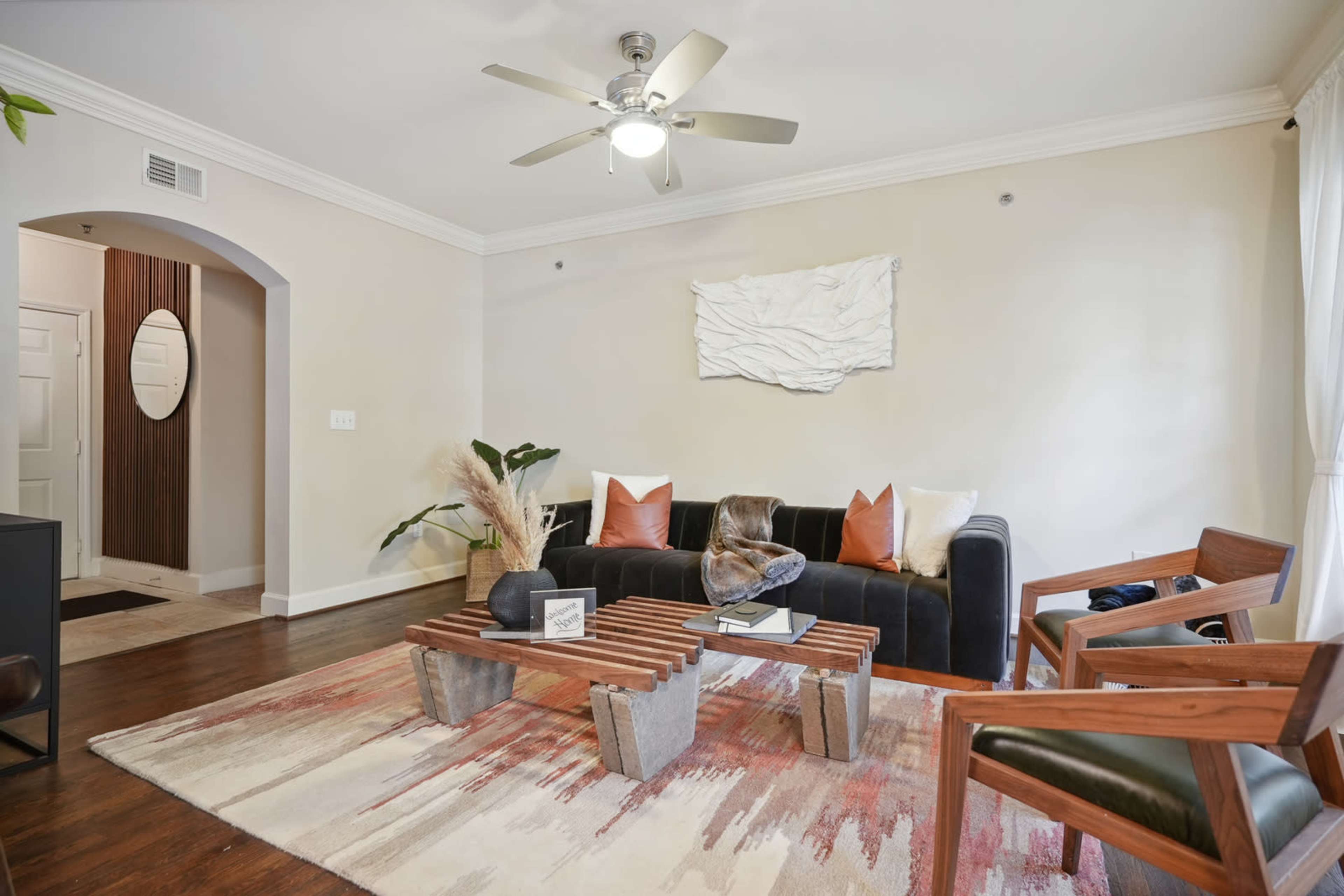The living room features a black sofa, a wooden coffee table, and two armchairs arranged on a patterned rug, with a ceiling fan above and a decorative wall piece.