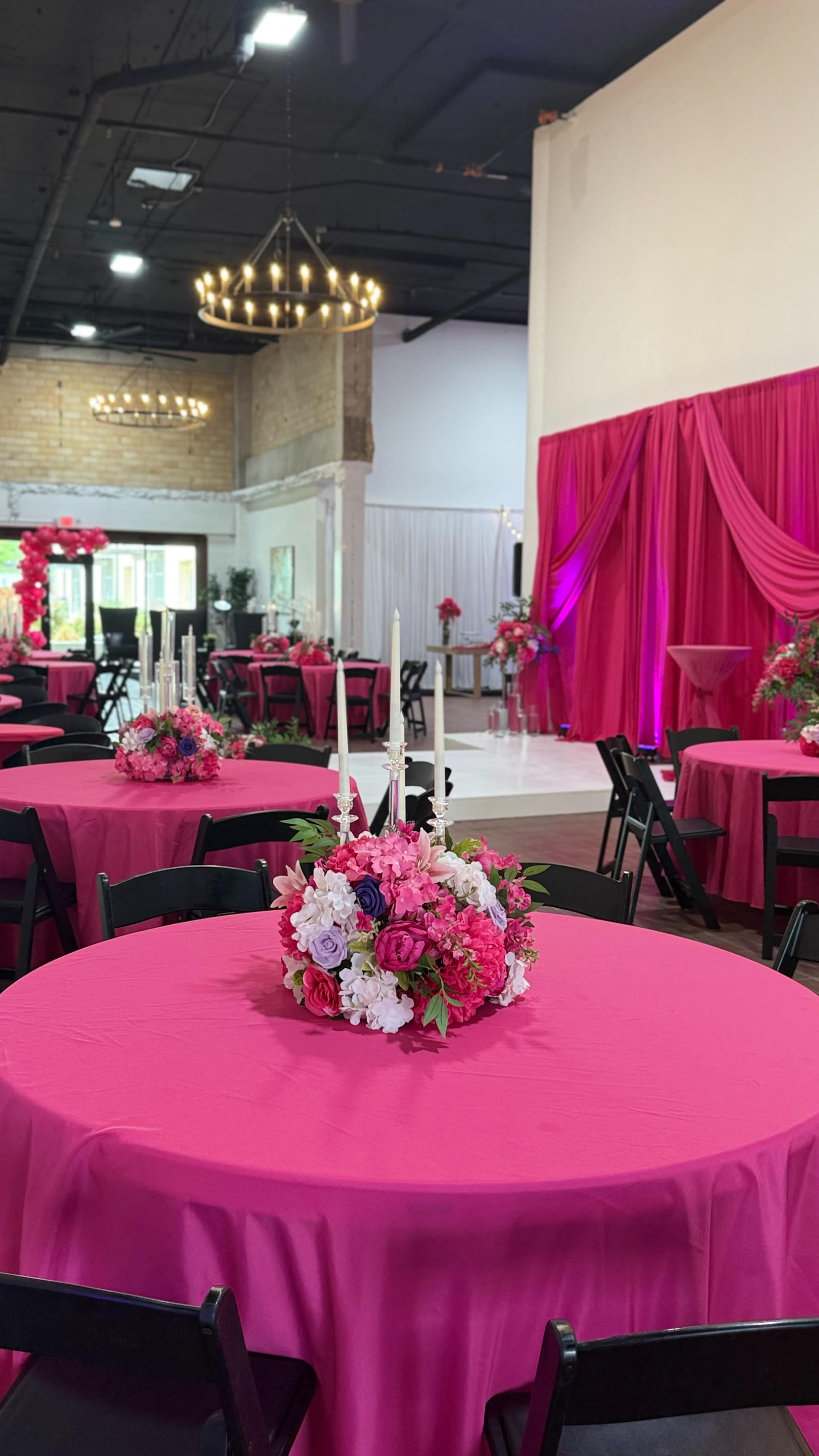 The room features round tables dressed in bright pink tablecloths, each adorned with floral centerpieces, against a backdrop of pink drapery.